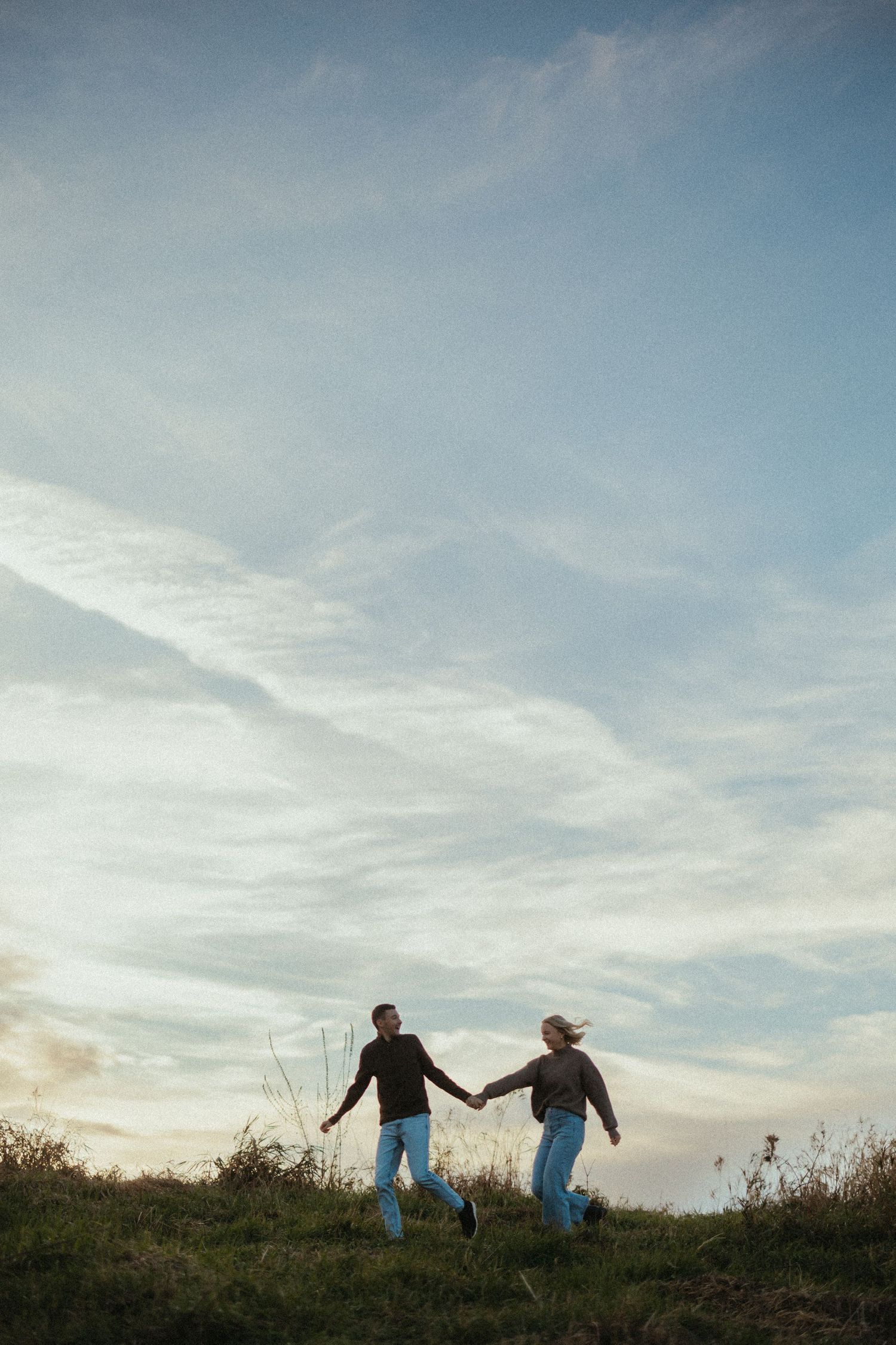 Two people hold hands while walking along a hilltop silhouetted against a cloudy sky.