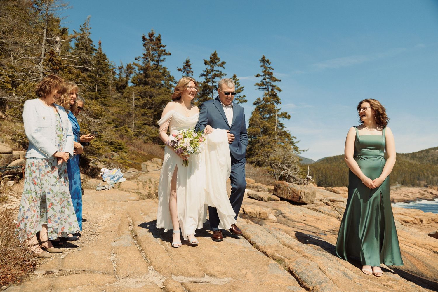 Bride in white dress walks with groom and wedding party on rocky coastal cliffs surrounded by pine trees on sunny day.