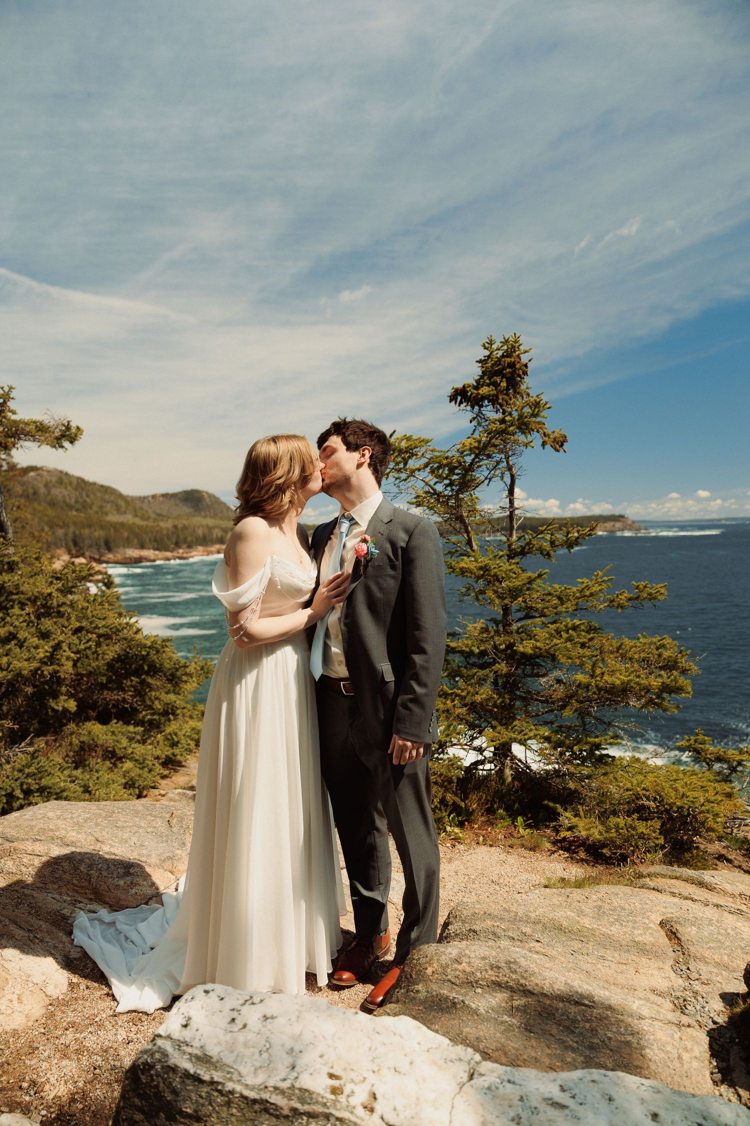 Bride and groom kissing on rocky coastal overlook, wearing wedding attire, with ocean and pine trees in background.