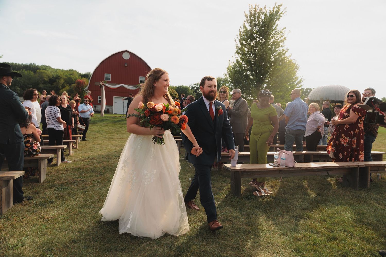 Outdoor wedding reception at sunset with guests mingling on a grassy hilltop overlooking scenic countryside views.