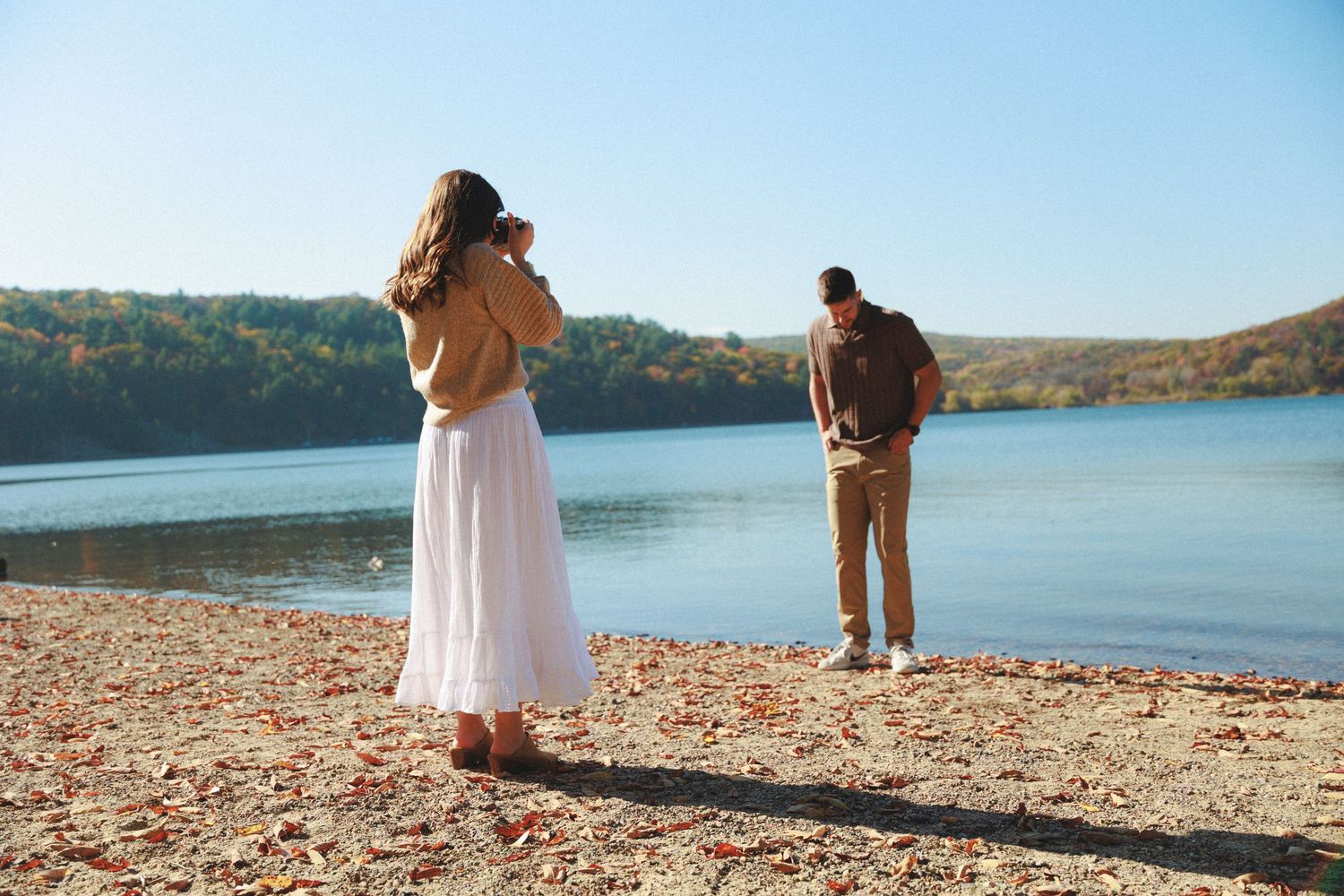 Couple taking photos by serene lake surrounded by fall foliage.