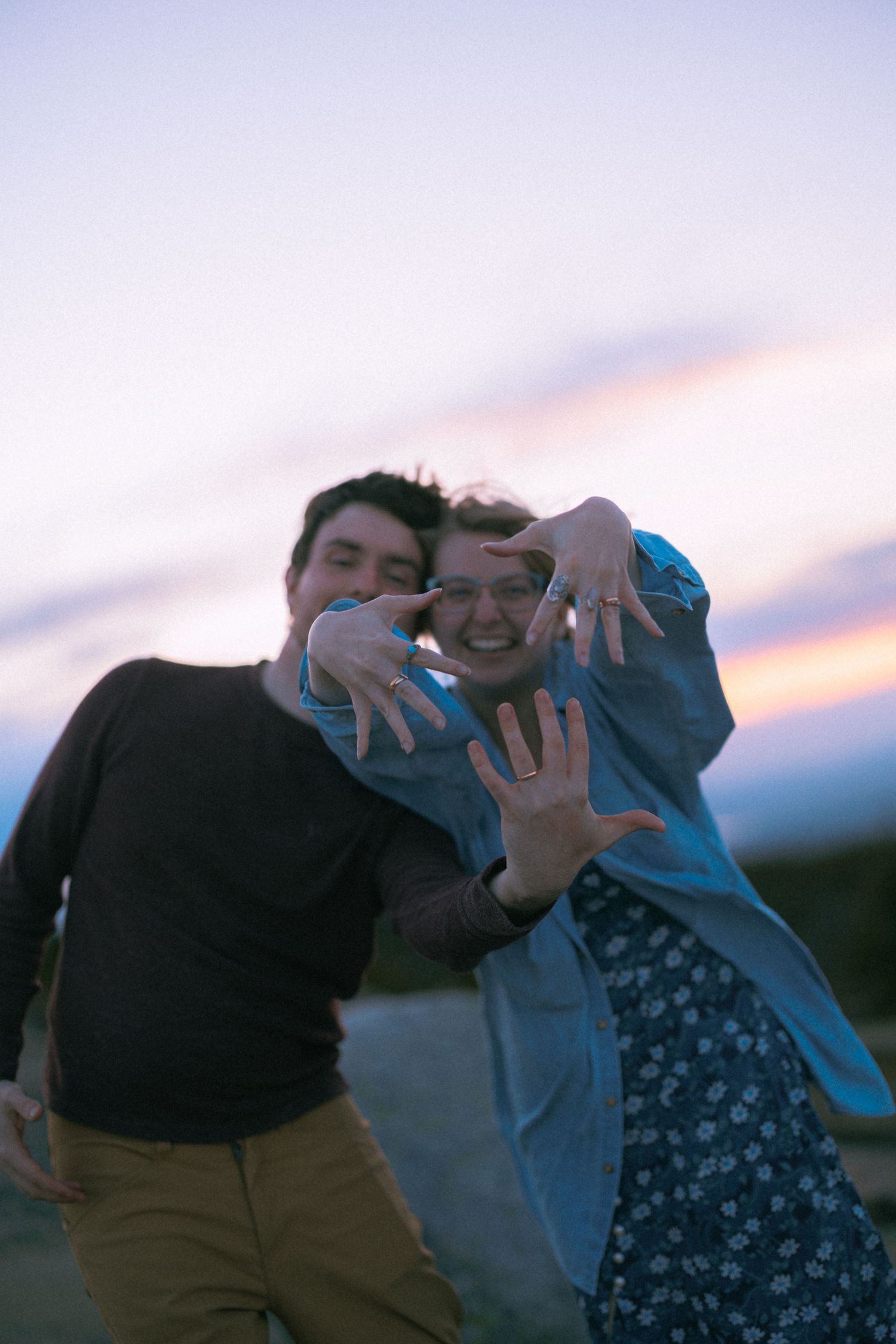 A joyful couple poses playfully at sunset, reaching hands toward camera against a colorful dusk sky.