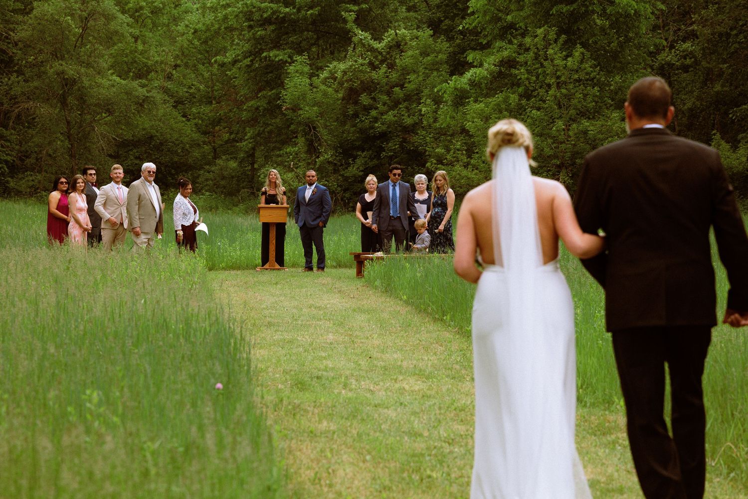 Wedding guests gather in a lush green field surrounded by trees.