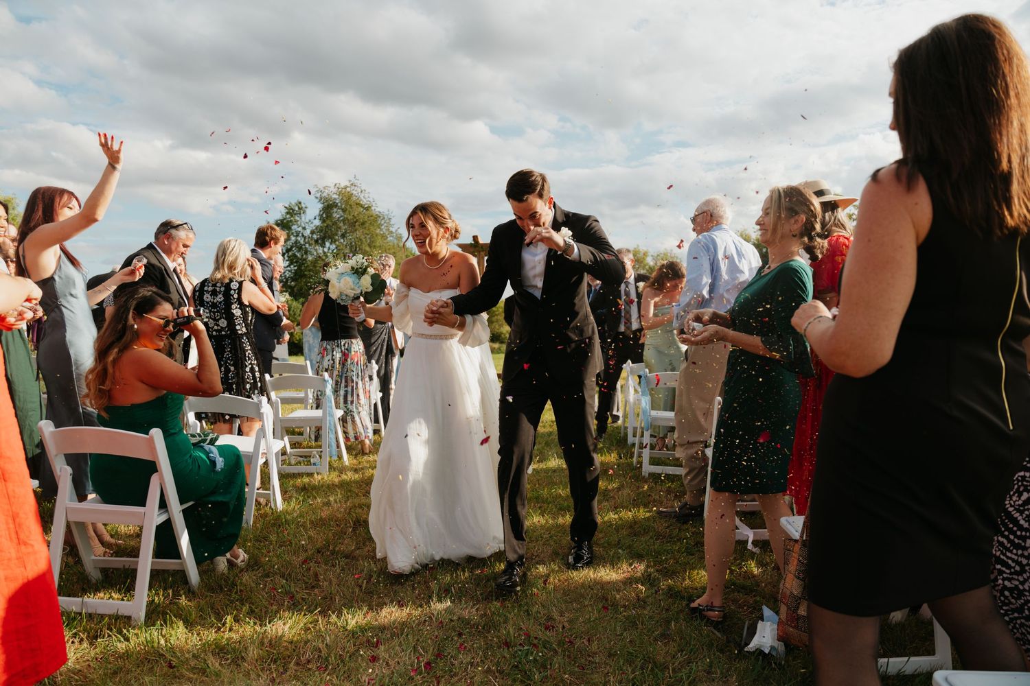 Wedding guests celebrate outdoors on a grassy hillside during a sunny afternoon.