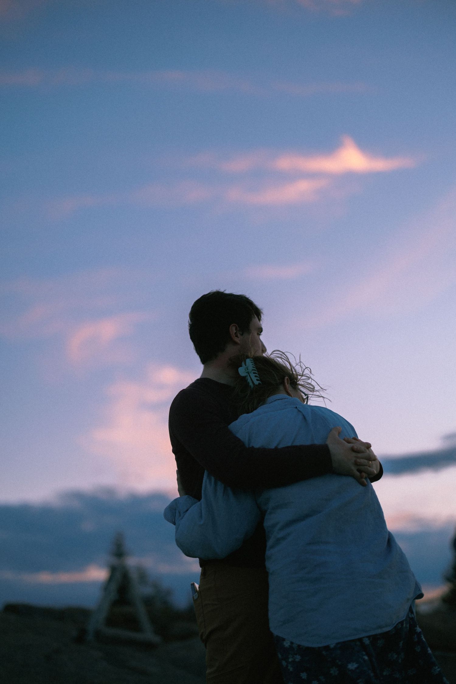 Couple embracing at dusk under a dramatic blue and pink cloudy sky, viewed from behind in a romantic outdoor setting.