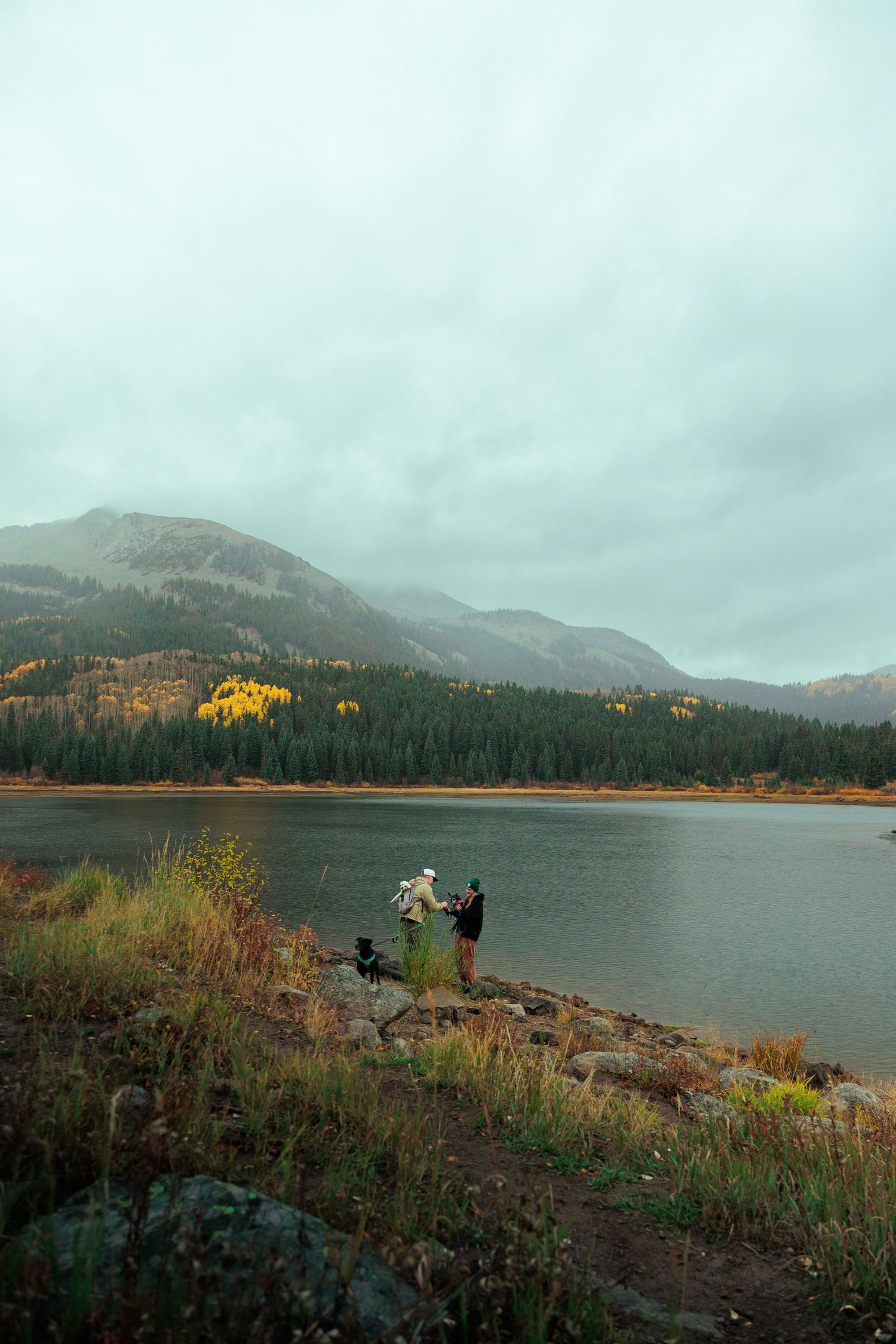 Two people stand by a mountain lake surrounded by autumn foliage and evergreen forests.