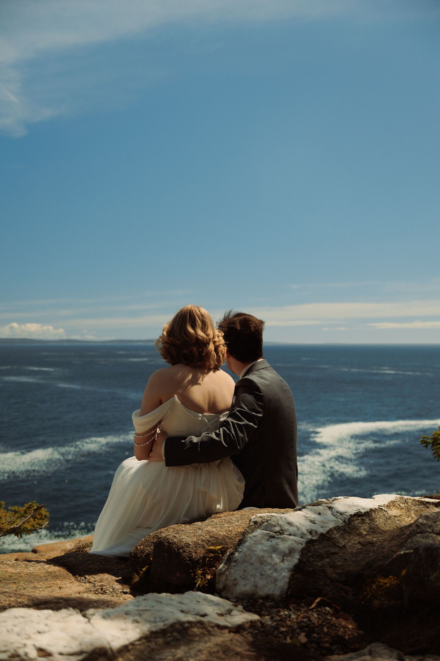 Bride and groom sit embracing on coastal rocks overlooking the ocean on their wedding day.