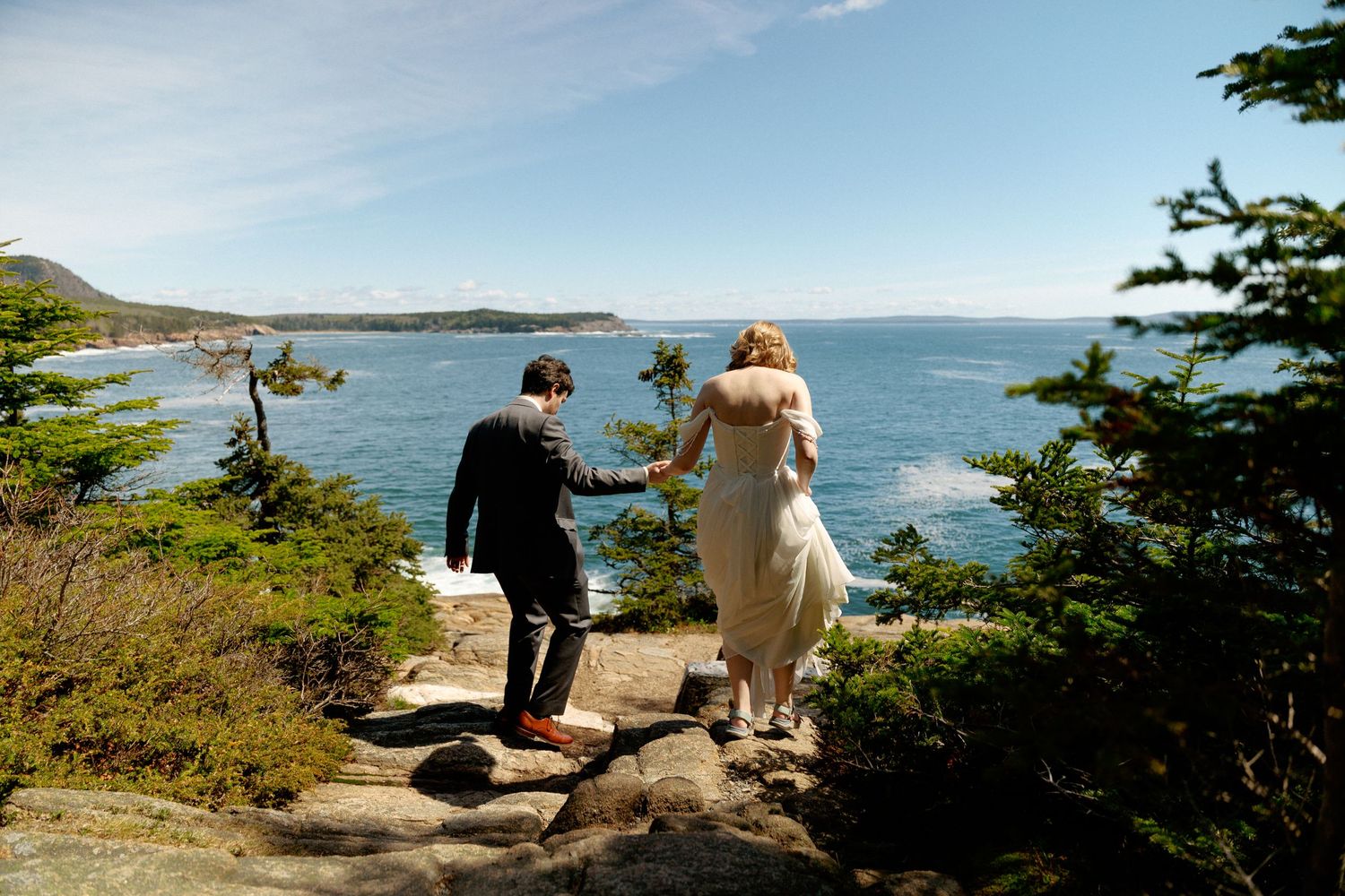 Bride and groom holding hands on rocky coastal overlook surrounded by pine trees with ocean views on their wedding day.