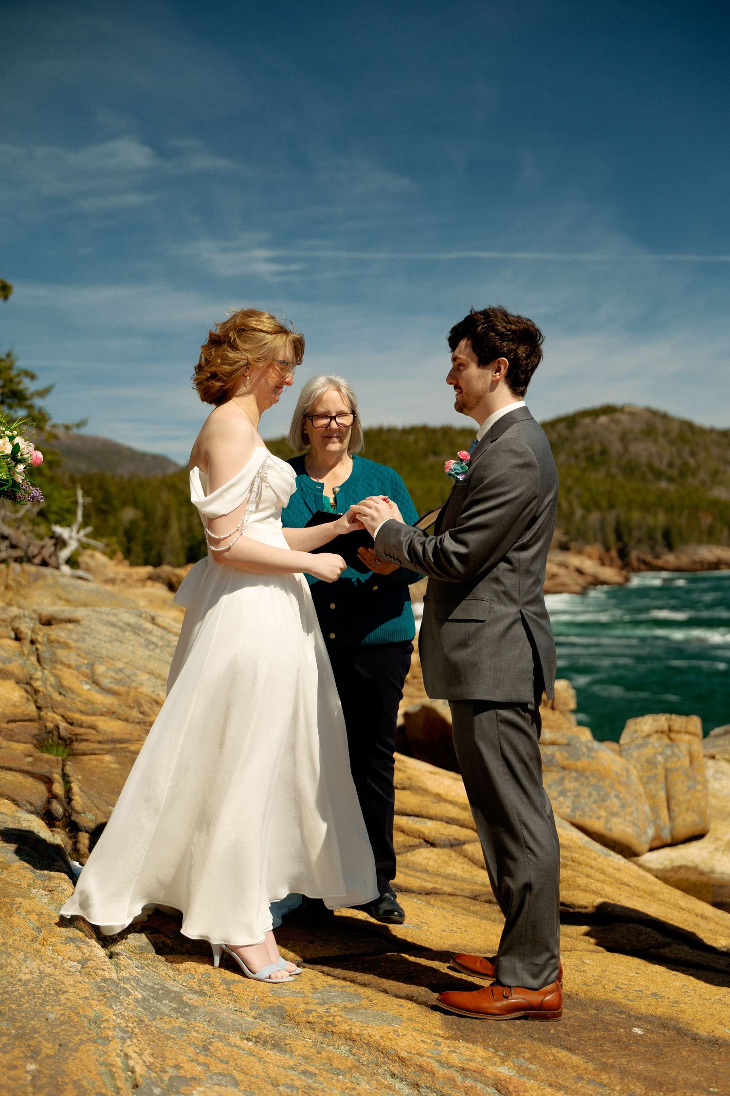 Couple exchanges vows on rocky coastal cliffs with officiant, ocean views, bride in white gown, groom in gray suit, sunny day.
