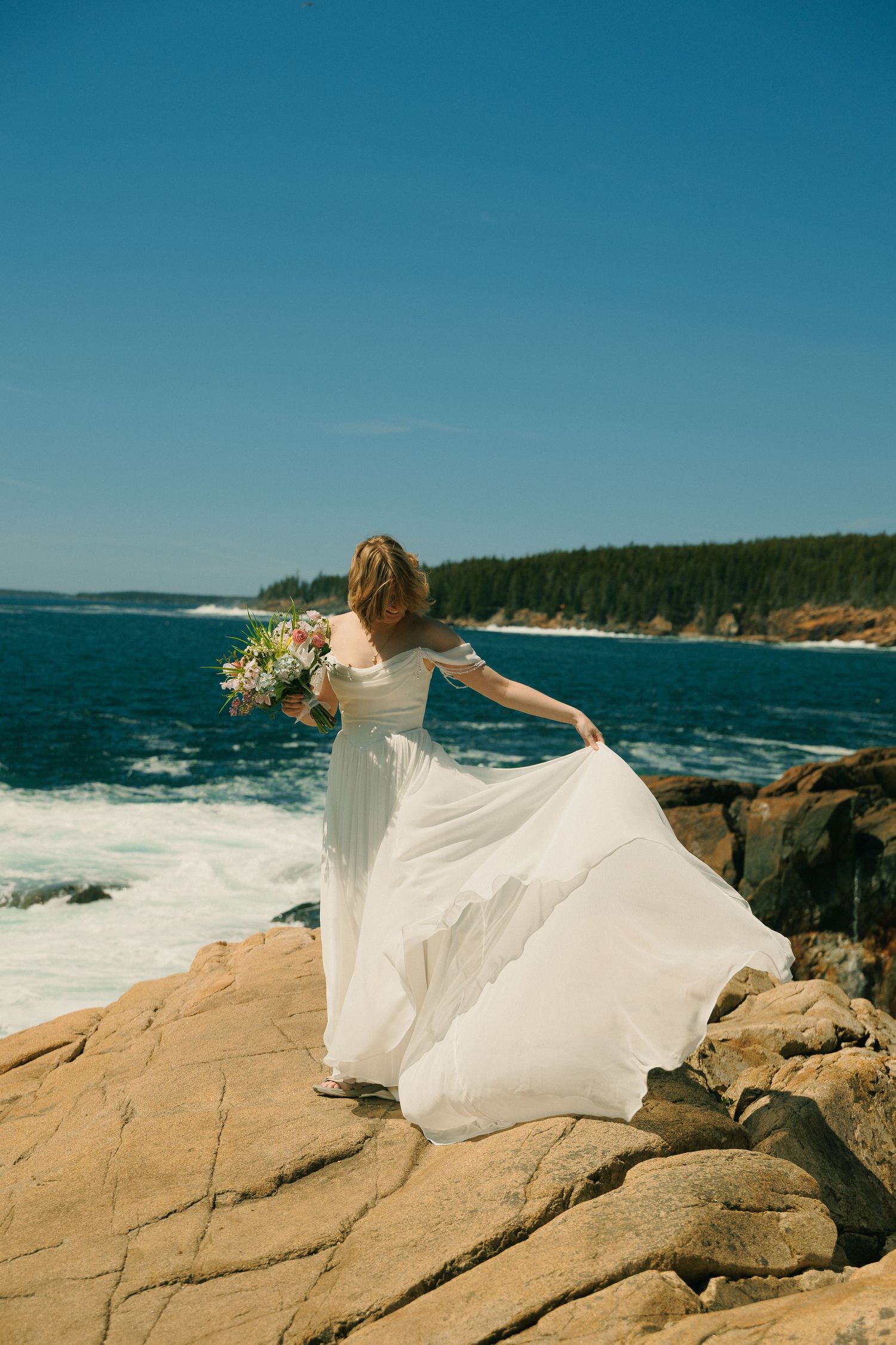 Bride in flowing white gown holds bouquet on rocky Maine coastline with waves below.