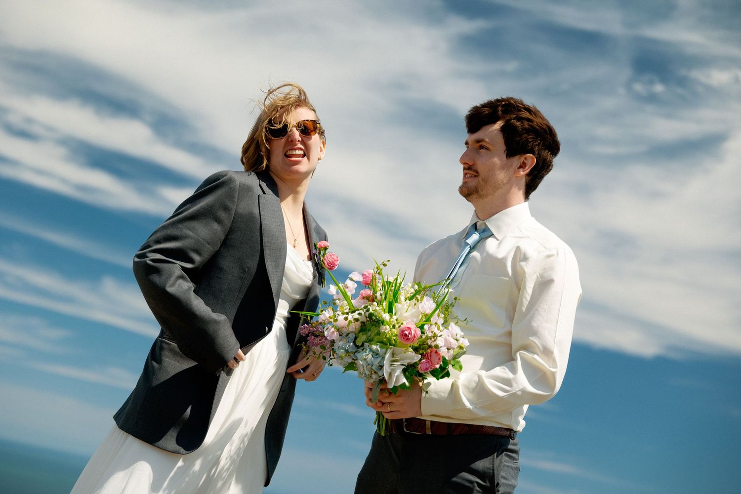 Smiling bride in sunglasses and groom holding colorful bouquet laughing together under a bright blue sky.