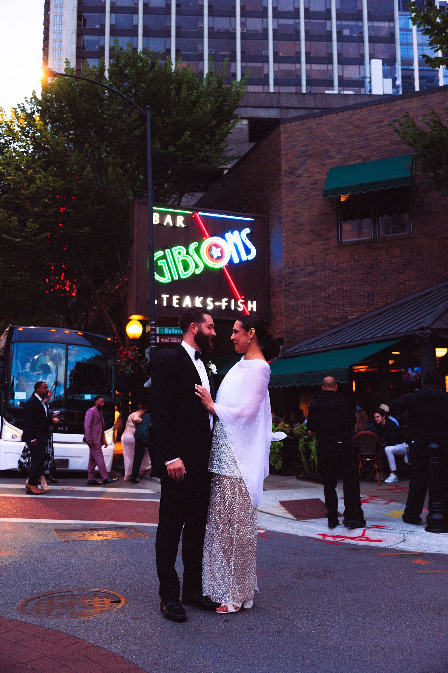 Wedding party gathered outside a brick building with neon signs at dusk.