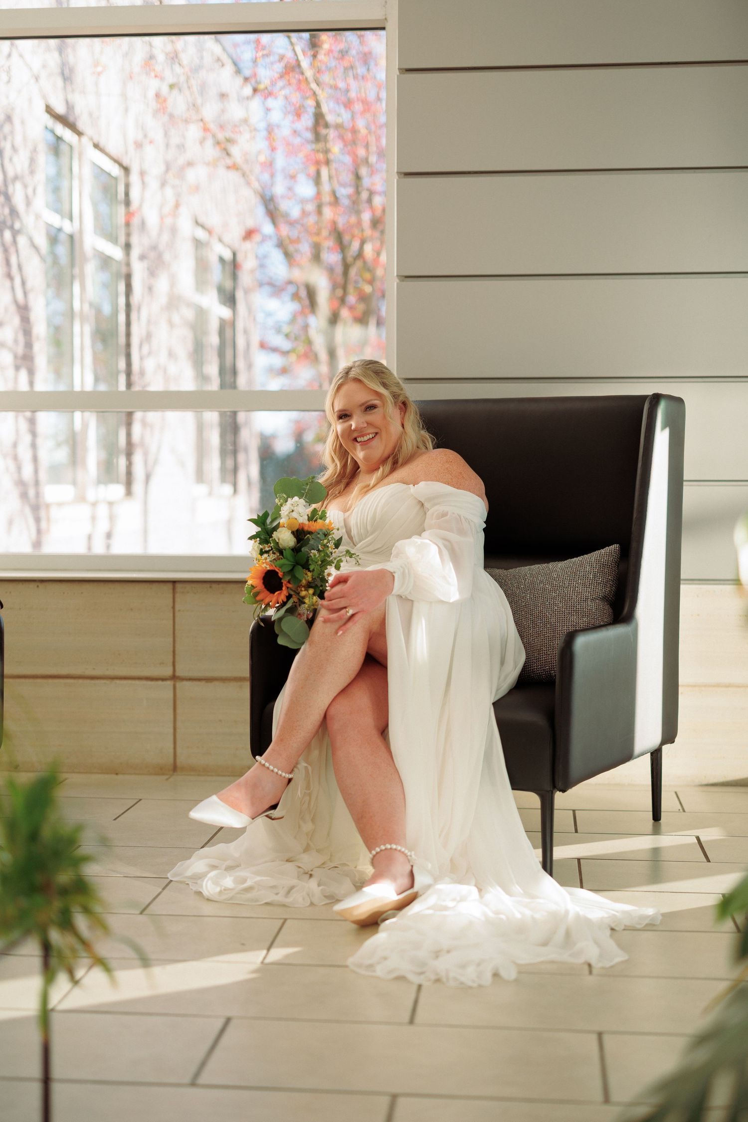 A bride in a white dress and pink shoes poses on a porch with autumn trees in the background.