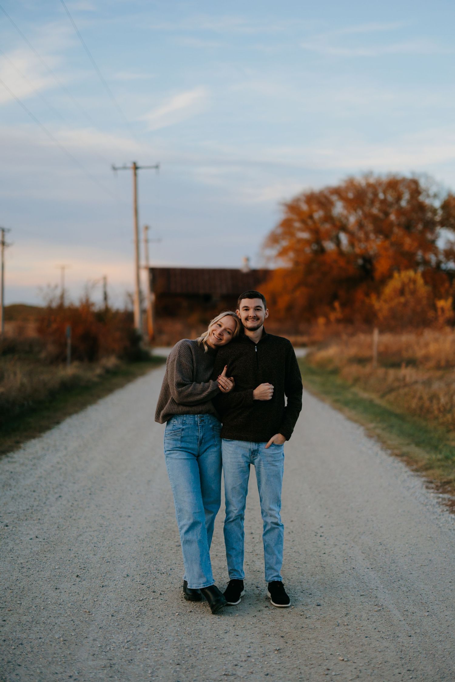 A couple walks together on a gravel country road during golden hour with autumn trees in the background.