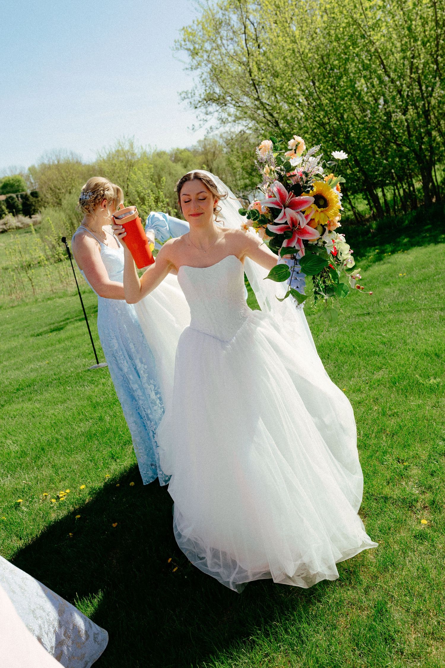 Bride in white ball gown holding colorful bouquet with sunflowers outdoors, assisted by bridesmaid on sunny day.