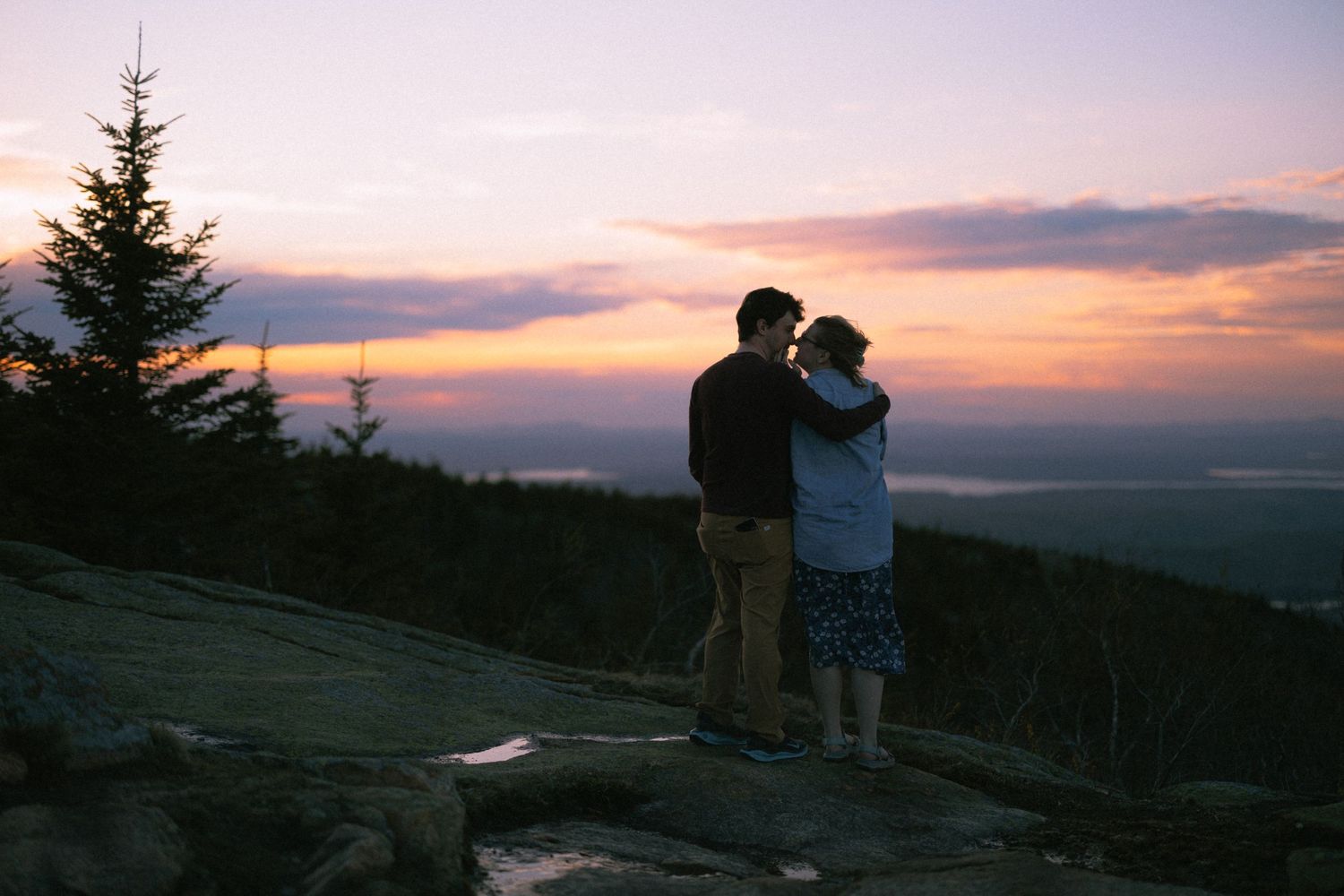 Couple embracing at sunset on a rocky mountain overlook with vibrant pink and orange sky.