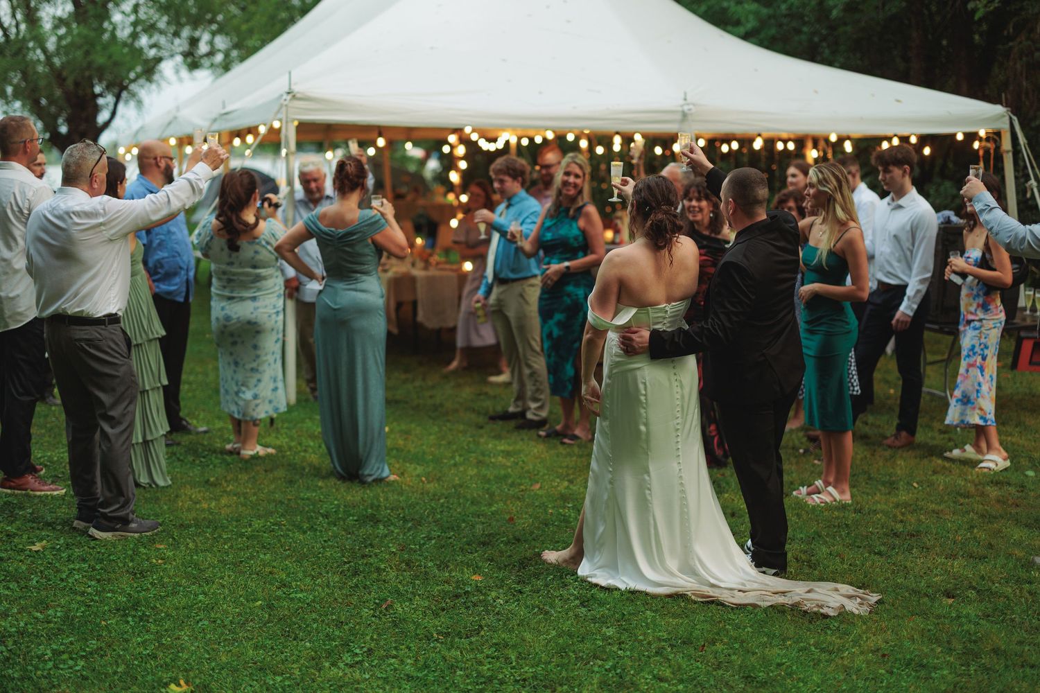 Wedding celebration under a white tent with guests gathered on a lawn.