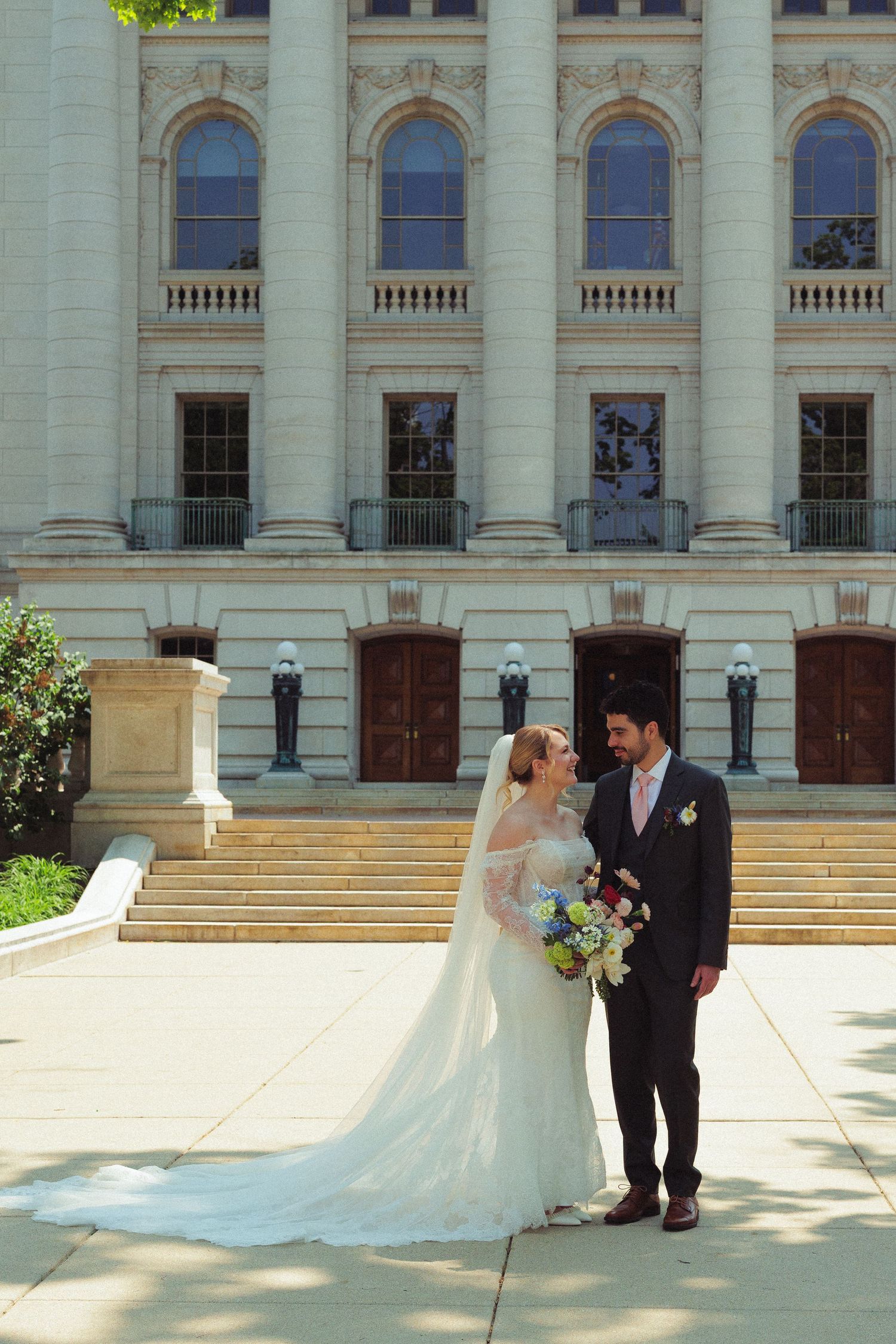 A wedding couple poses for photos on the grand steps of a historic building with classical architecture.