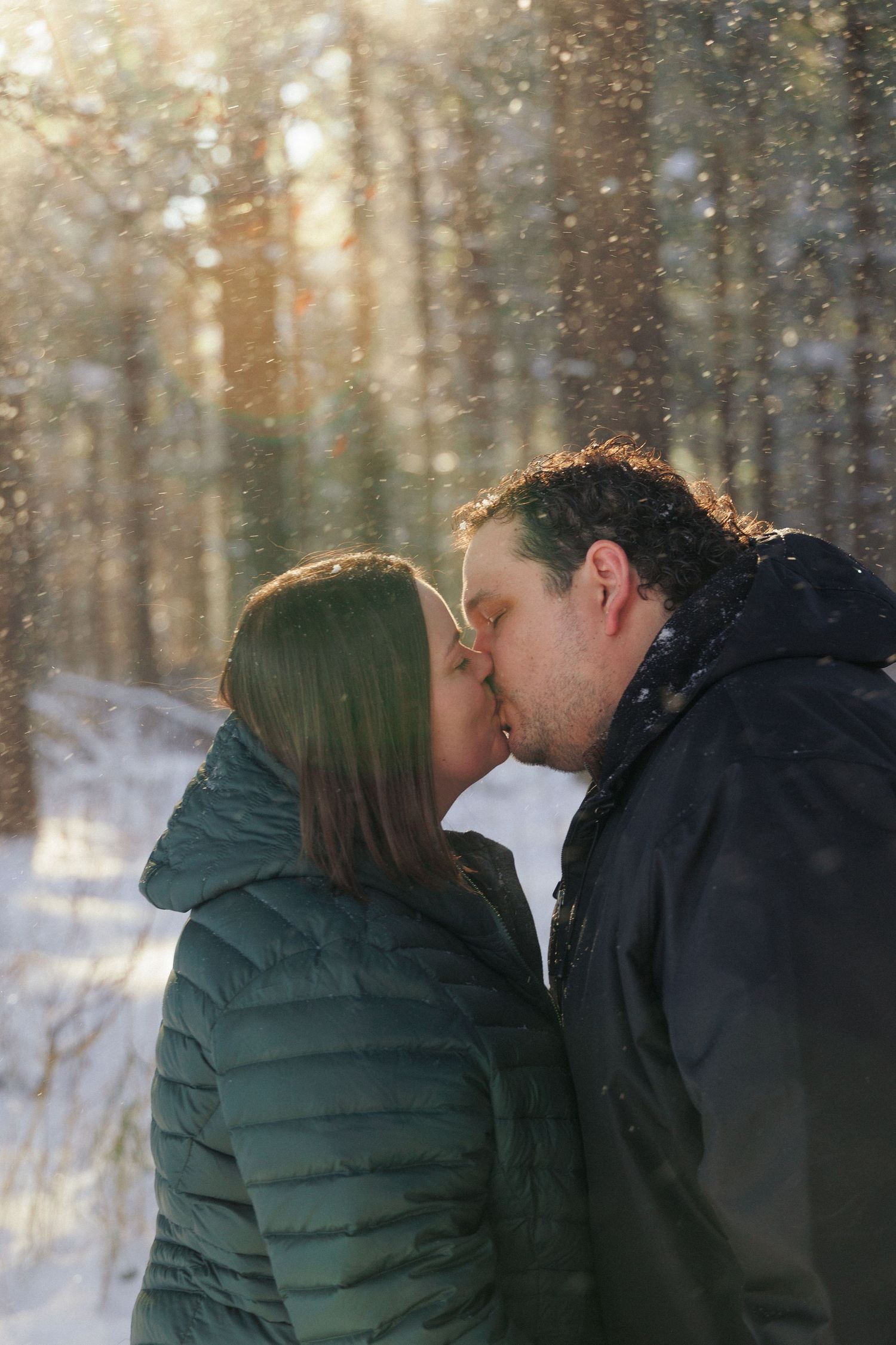 Intimate winter moment between couple in a snow-covered forest with sunlight streaming through trees.
