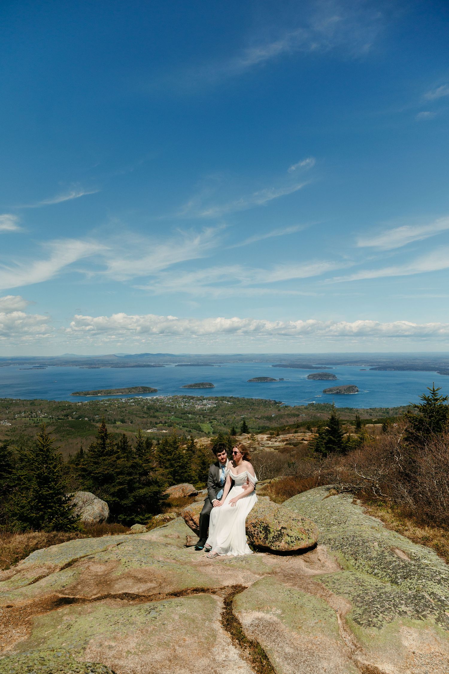 Bride and groom sit together on mountain summit with stunning coastal island views.