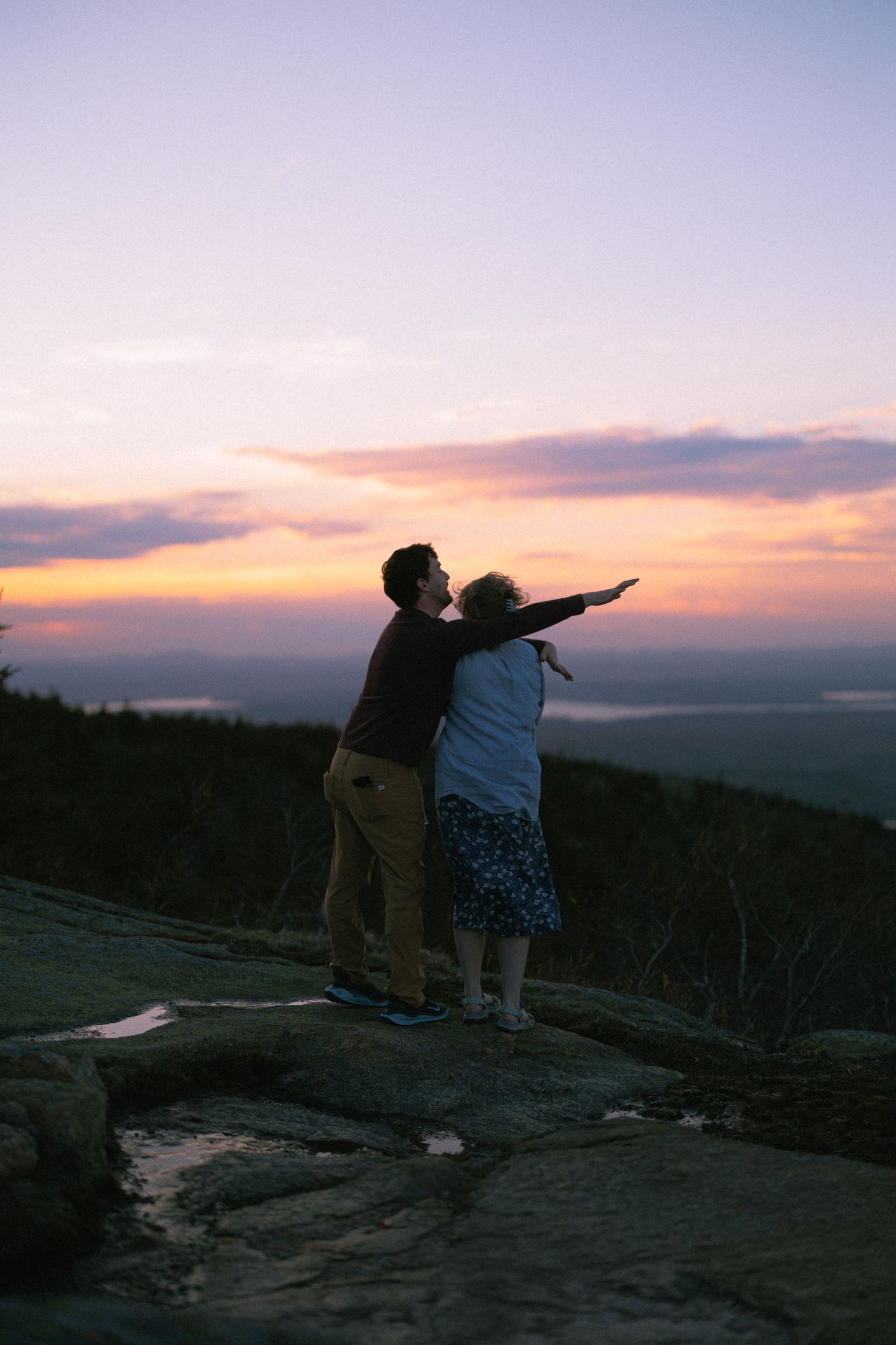 Couple standing on rocky summit embracing at sunset, pointing toward colorful orange and purple sky over mountain landscape.