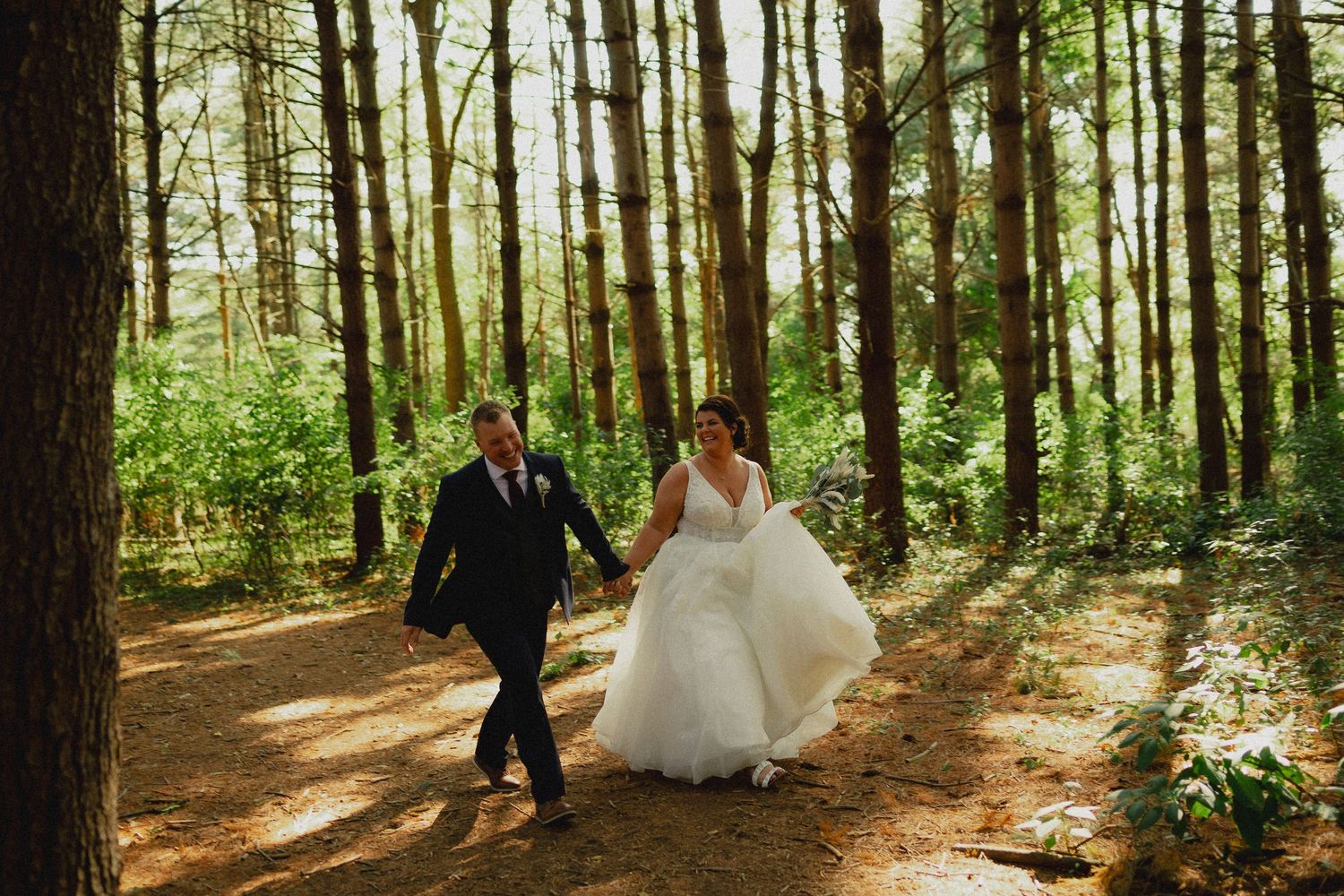 A couple in wedding attire walks hand in hand along a sunlit forest path surrounded by tall pine trees.