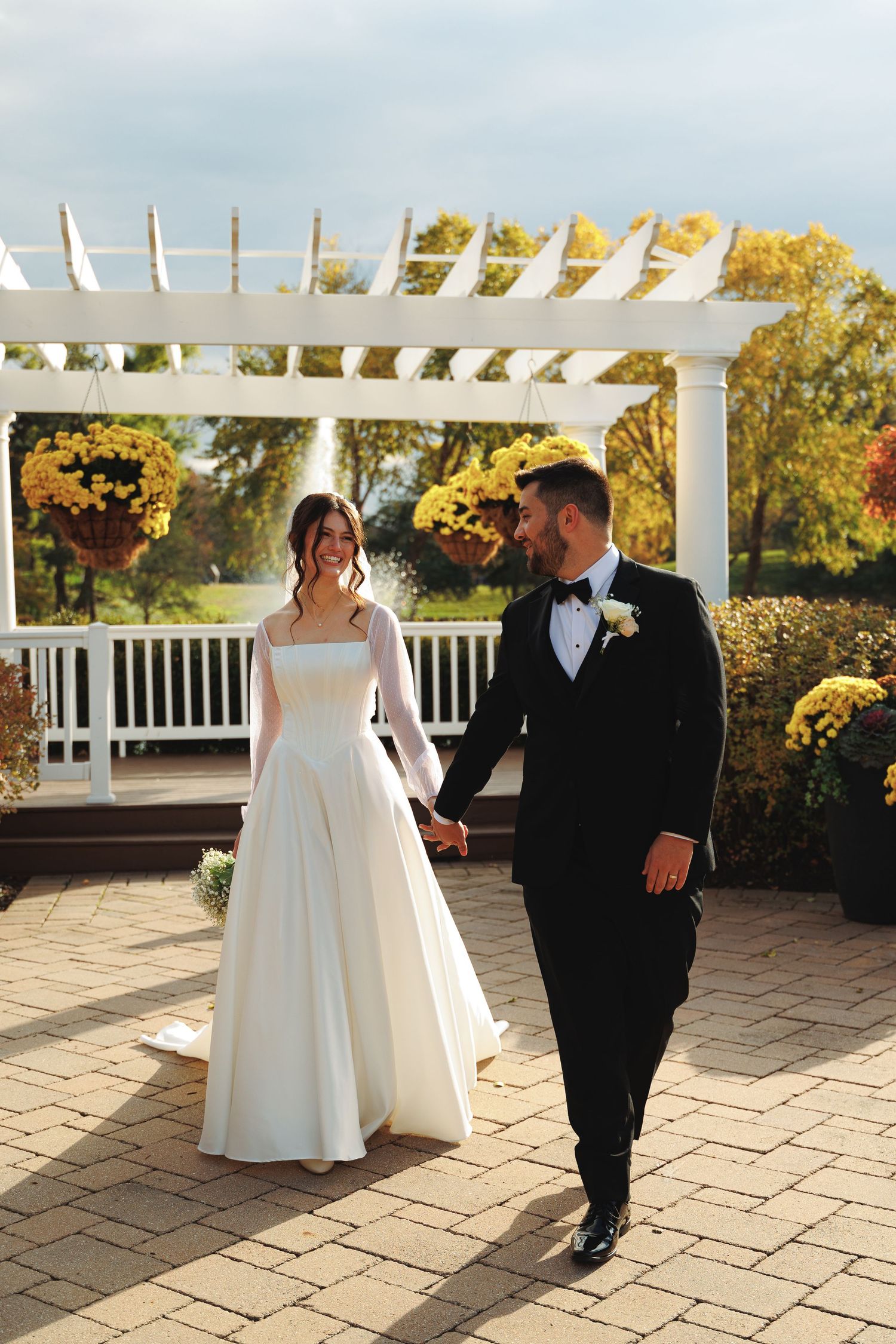 Couple walking together on stone patio under white pergola during autumn wedding.