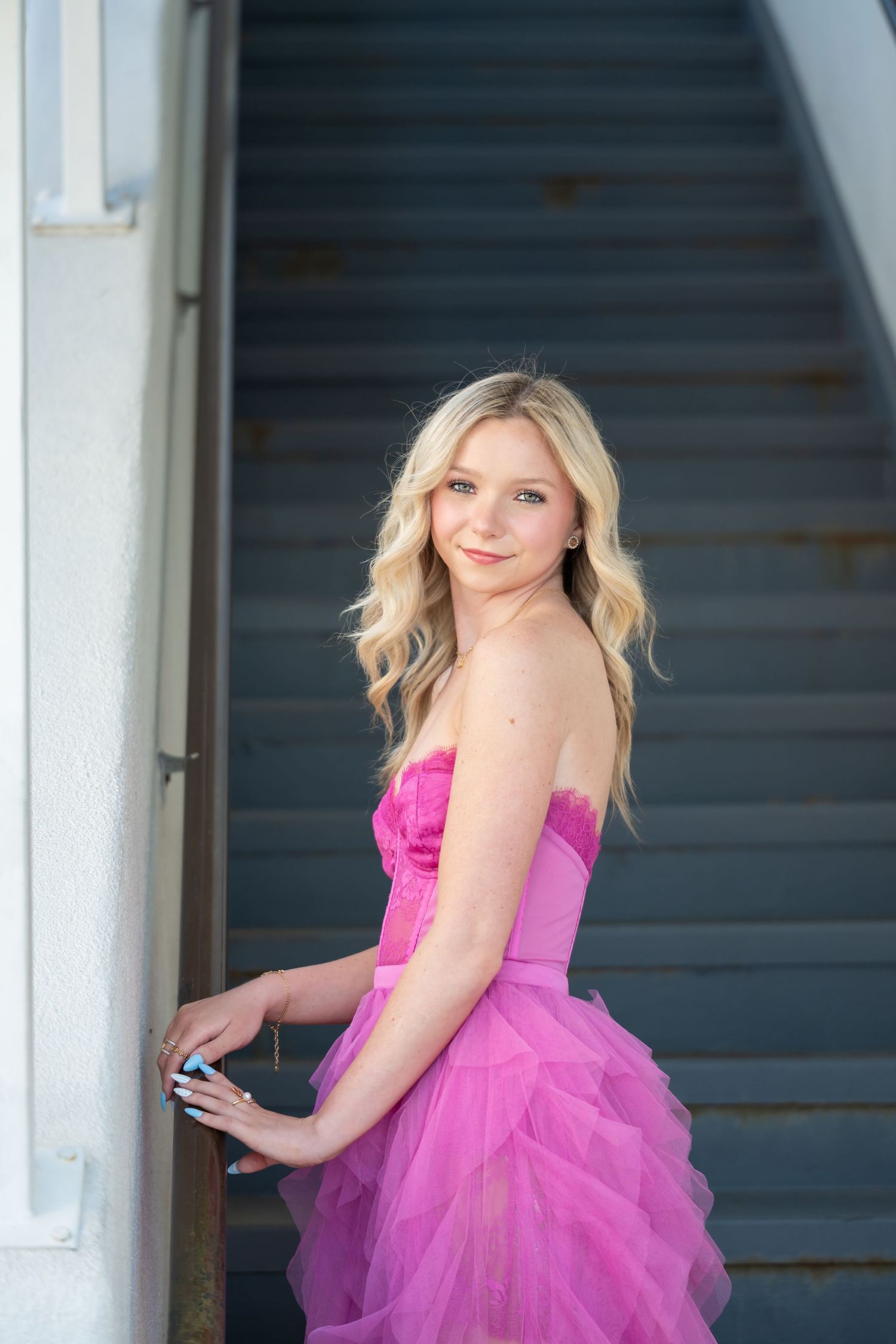 Young blonde woman in a strapless pink tulle dress poses gracefully on outdoor stairs.