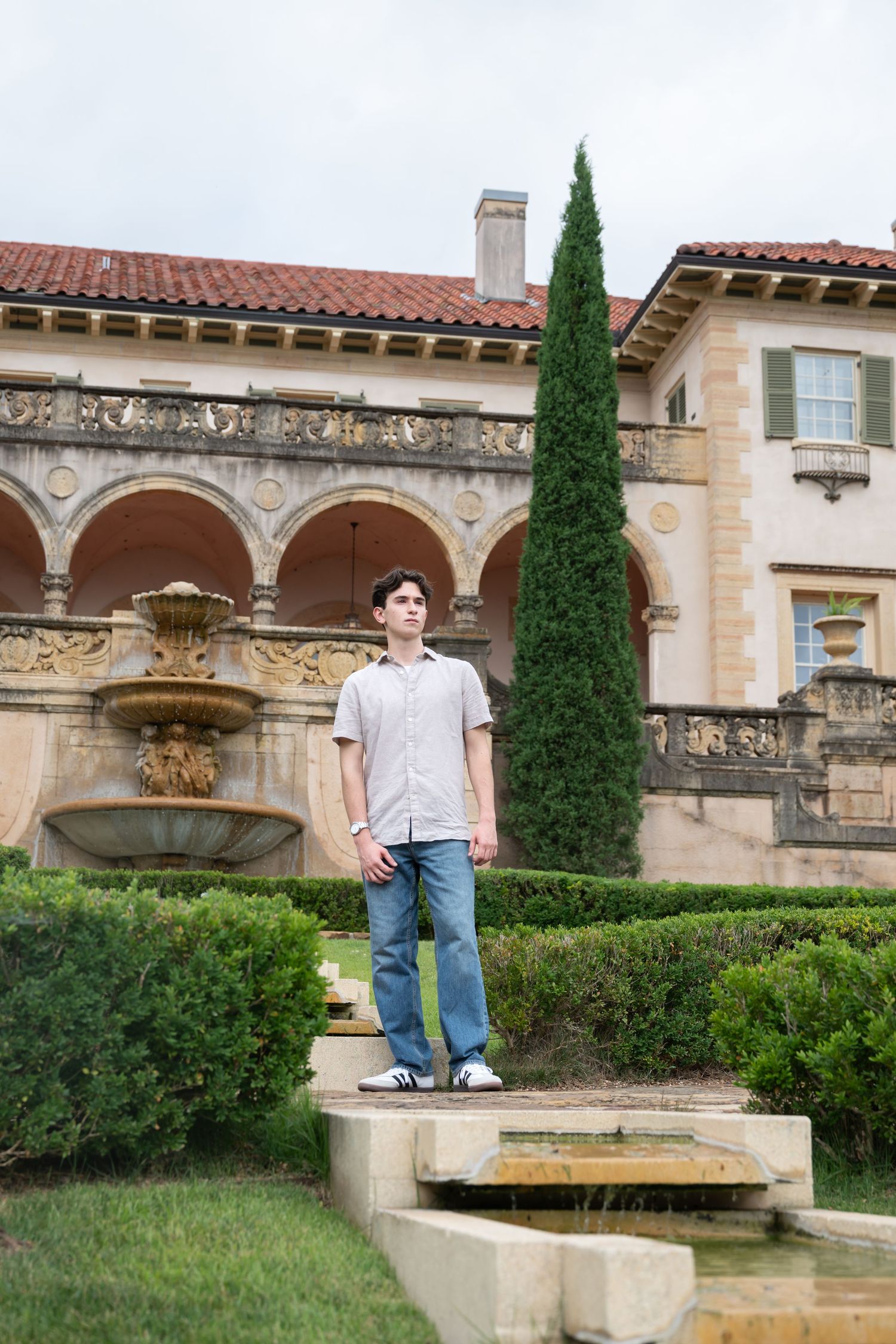 Young man standing on garden steps before an ornate Italian Renaissance-style villa with fountain and cypress tree.