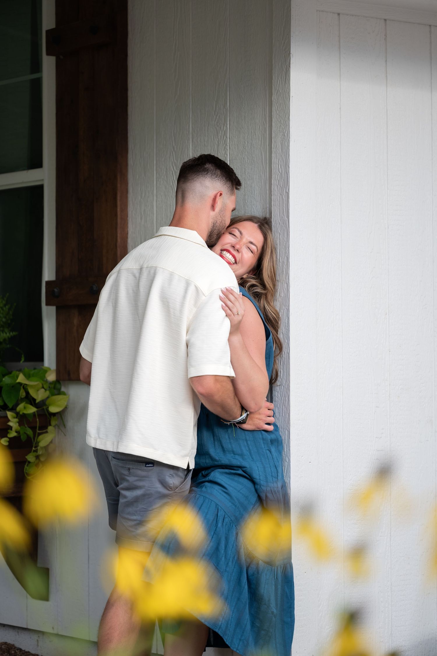 A couple embraces and smiles on a white porch, woman in teal dress, man in white shirt, yellow flowers in foreground.