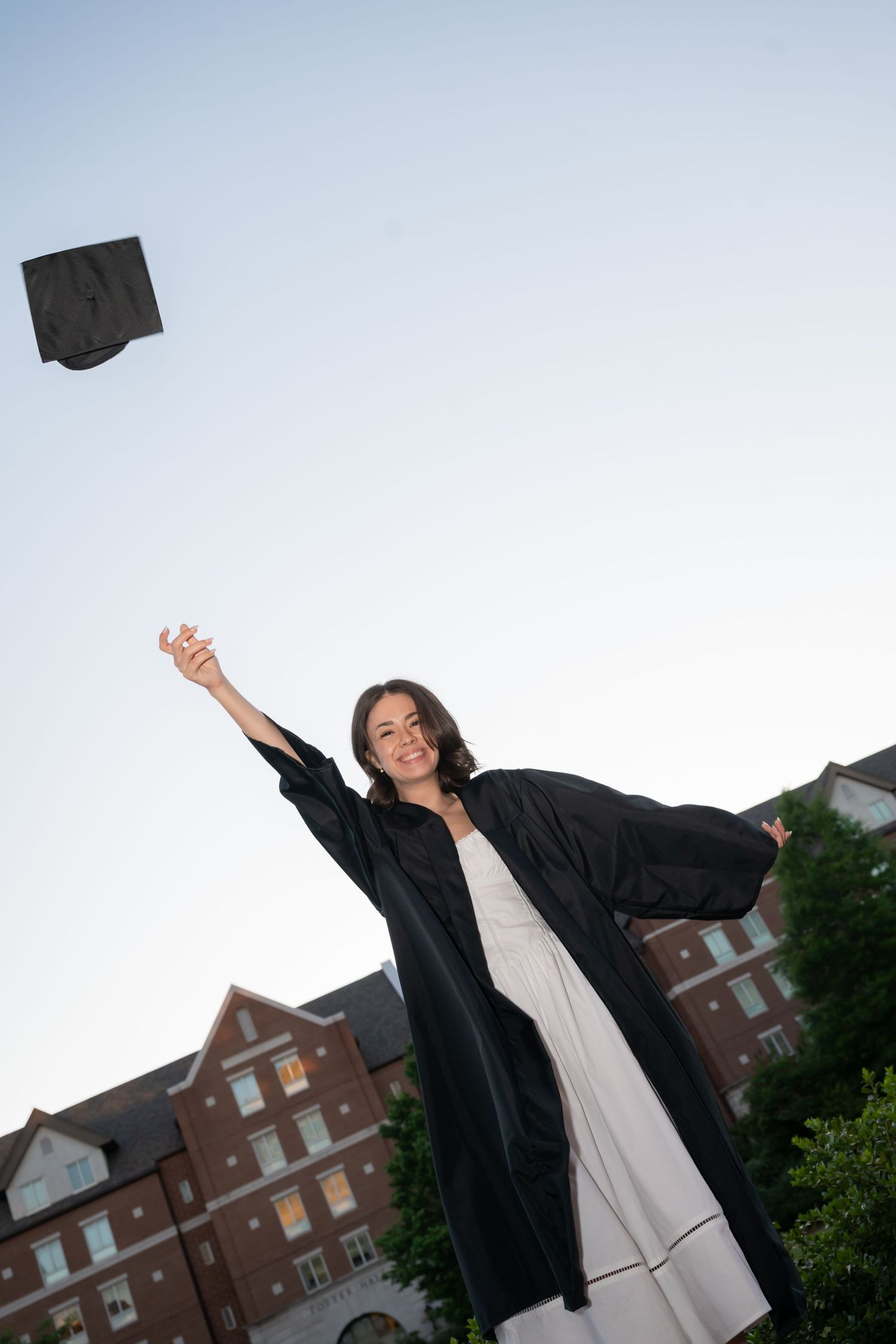 A smiling graduate in black gown tosses her cap in the air outdoors near brick buildings.