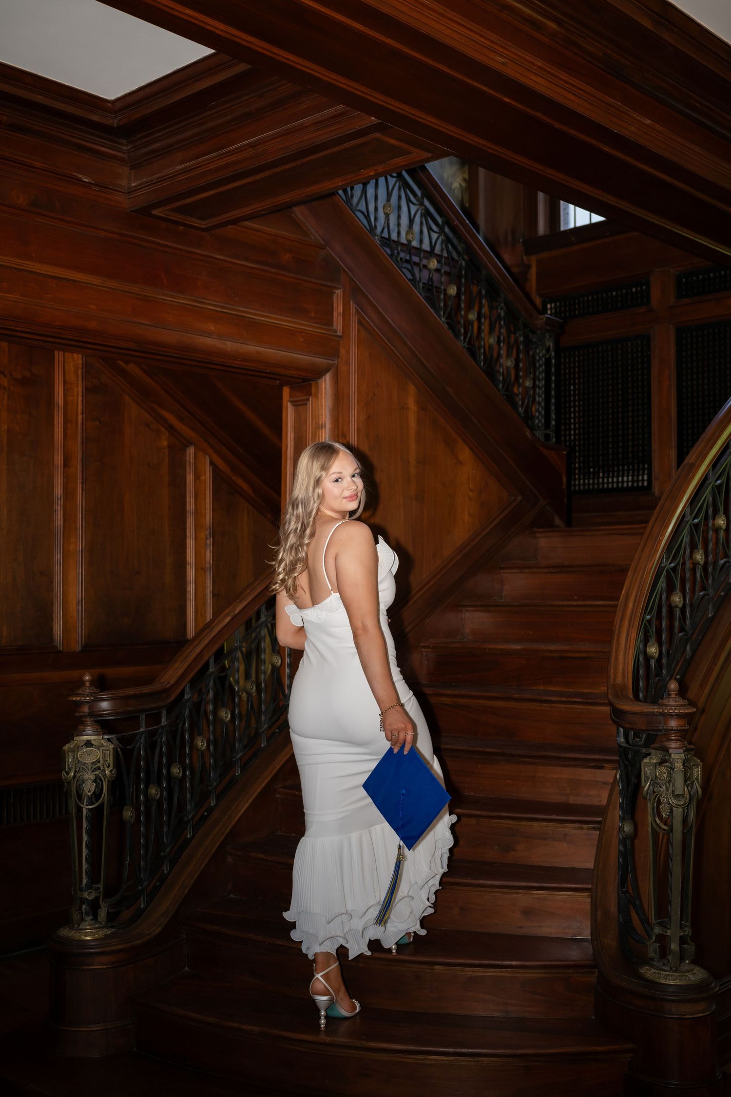 Woman in white ruffle dress holding blue clutch poses on ornate wooden staircase in elegant mansion.