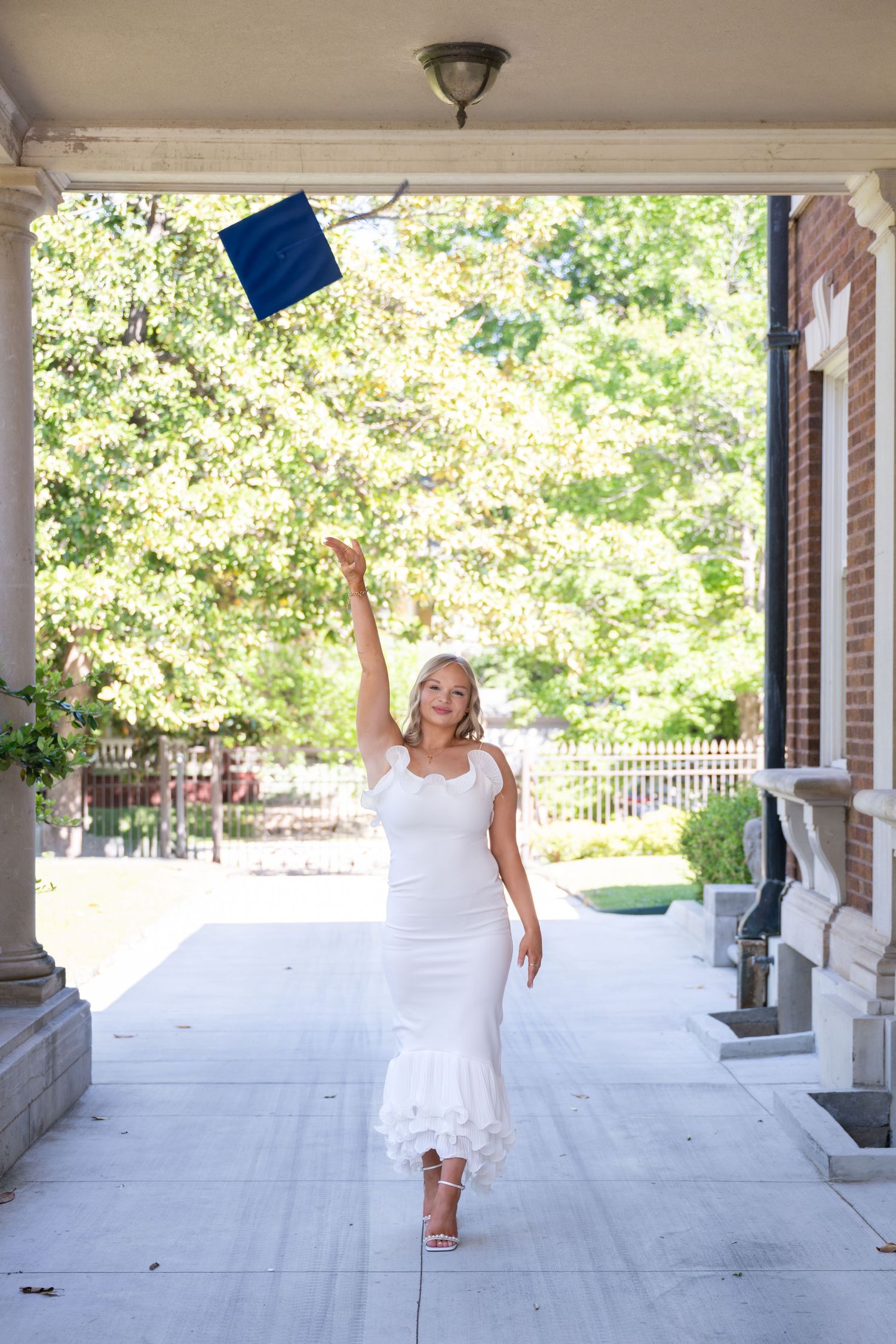 Woman in white dress tossing graduation cap in the air outdoors near brick building.