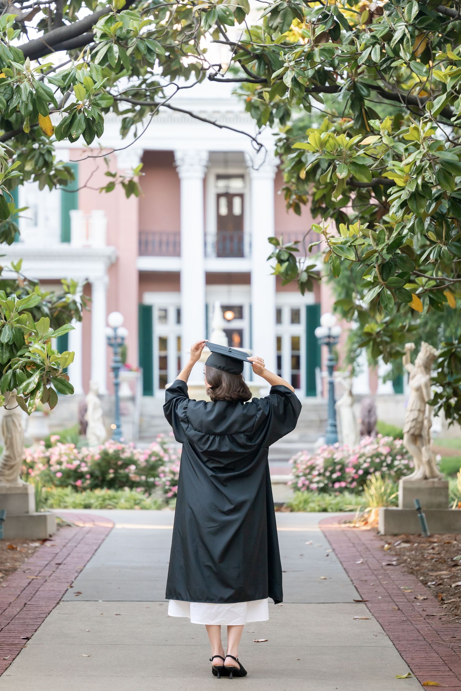 Graduate in black cap and gown stands on pathway facing elegant pink colonial building surrounded by lush green trees.