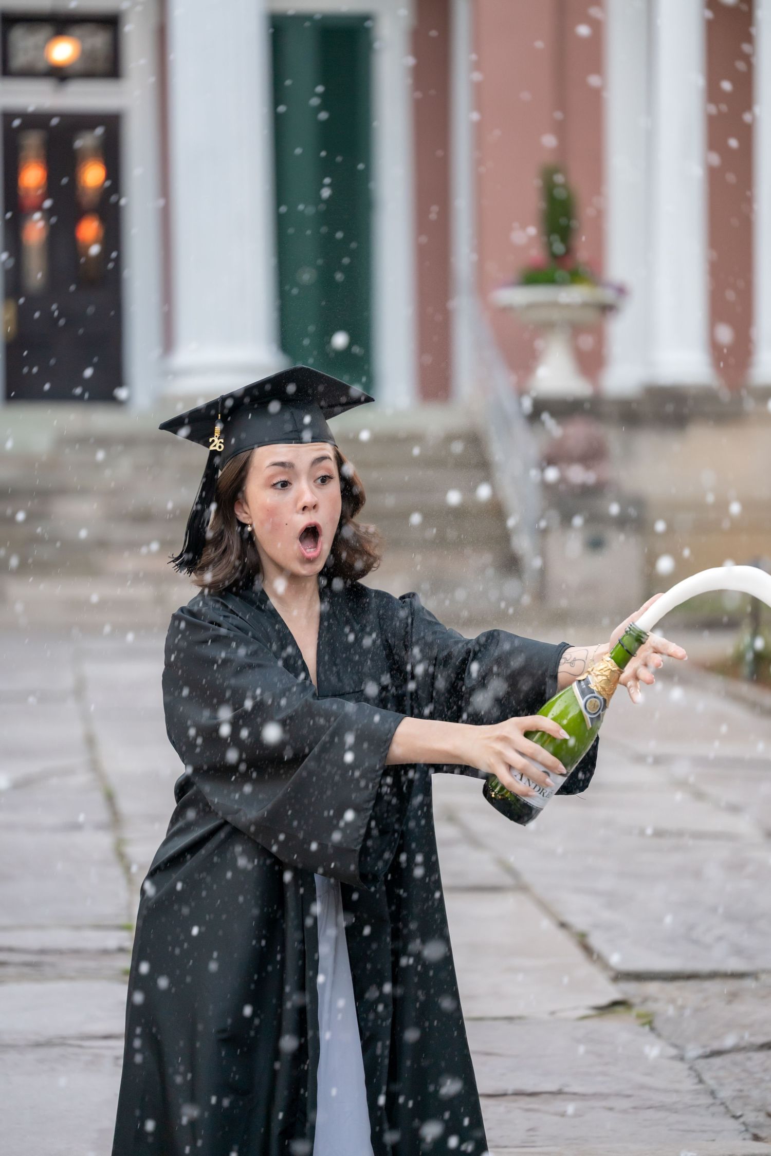 Graduate in black cap and gown pops champagne bottle outdoors in falling snow, looking surprised.