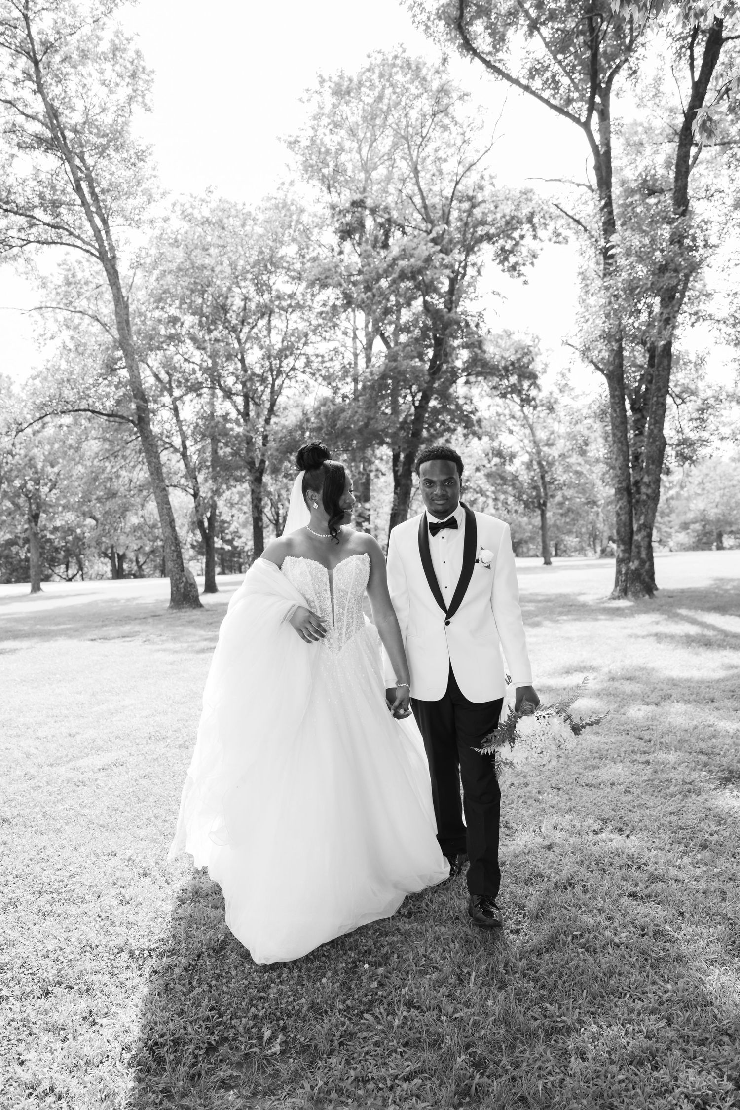 Black and white photo of a bride and groom walking hand in hand through a tree-lined park on their wedding day.