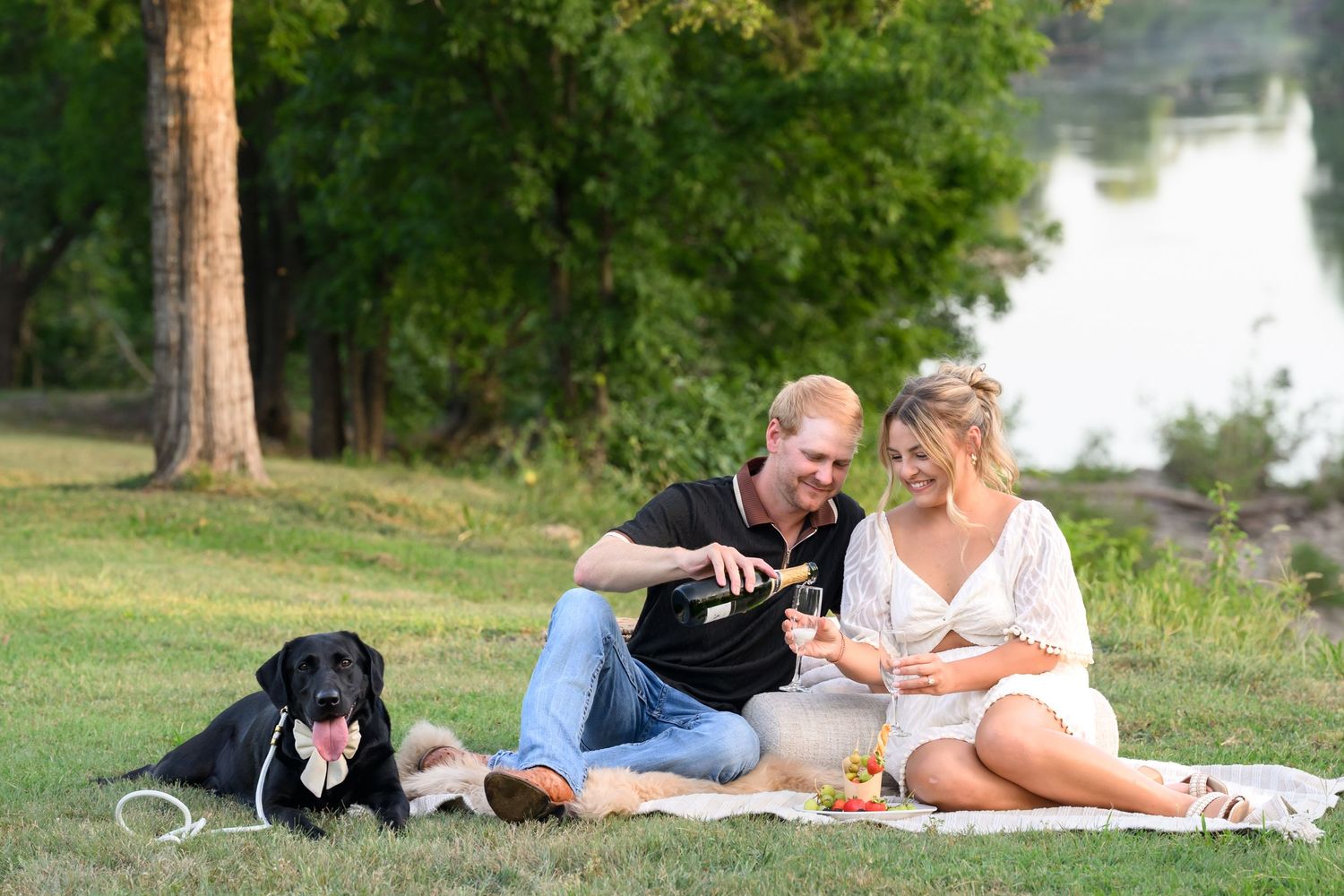 Couple sitting on blanket in park pouring champagne with black Labrador dog beside them near a lake.