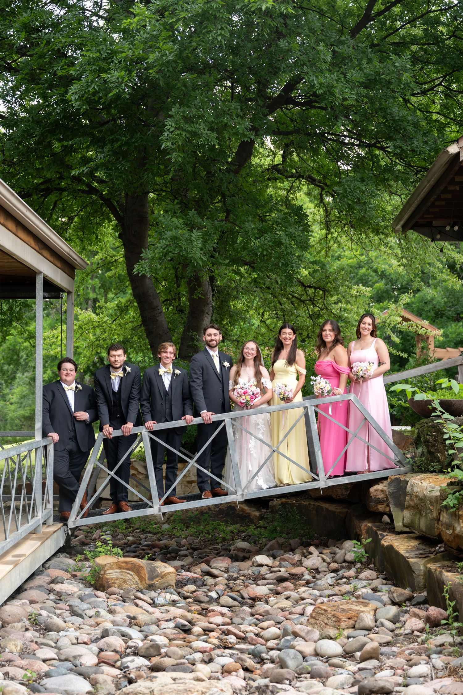 Wedding party posing on a small bridge surrounded by lush green trees and rocky stream at outdoor venue.