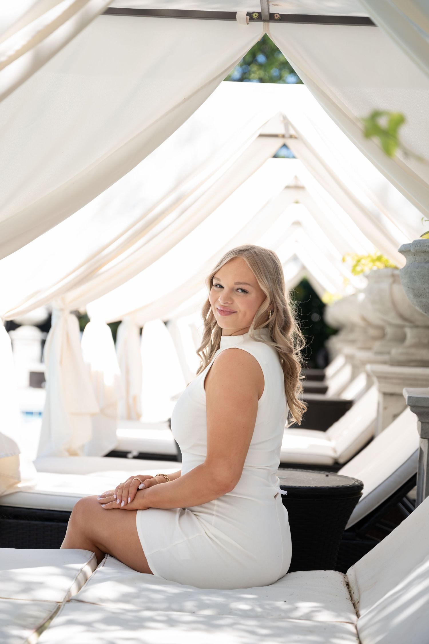 A blonde woman in a white mini dress sits elegantly on a luxury poolside daybed under a white cabana.