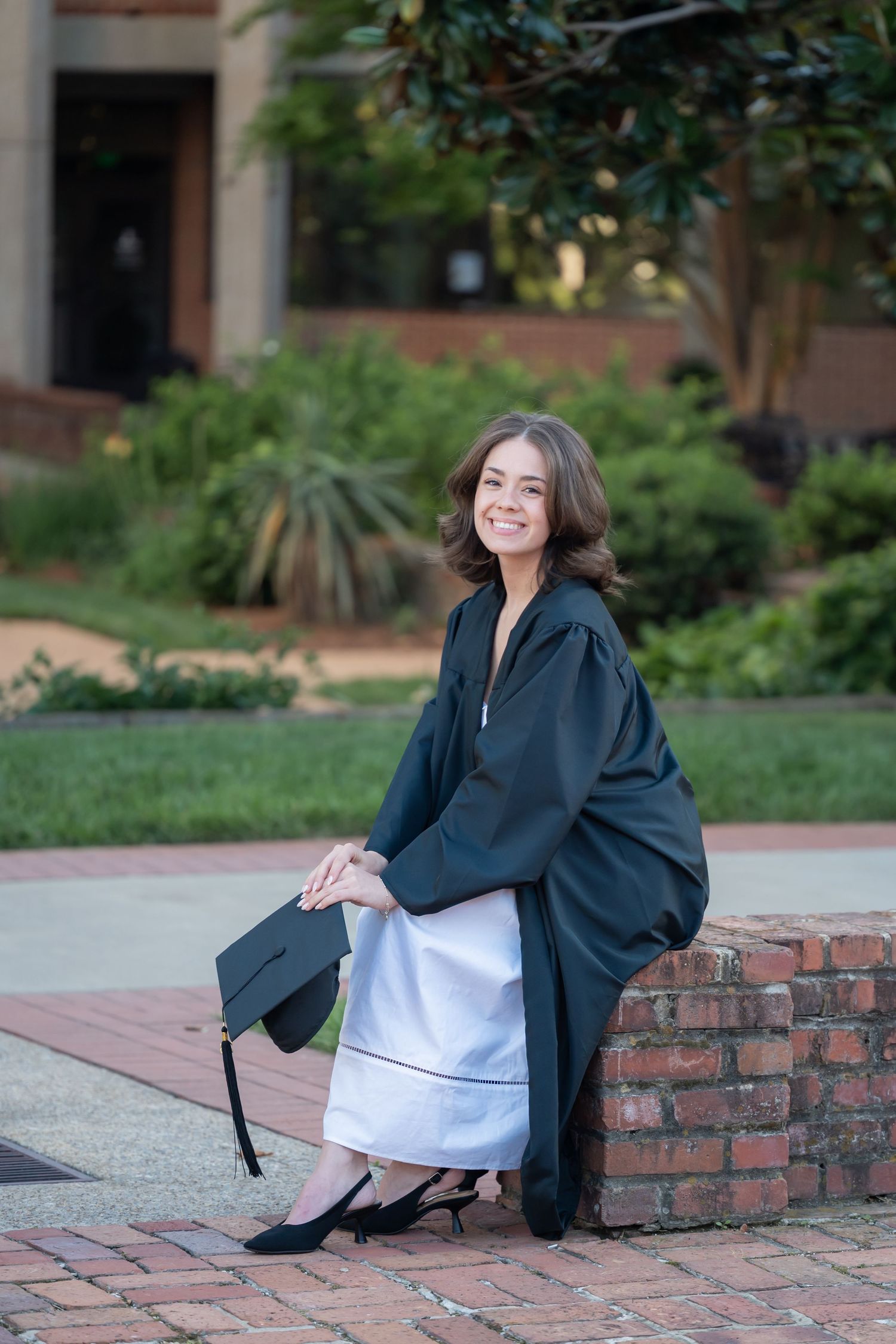 Smiling graduate in black gown and cap sitting on brick wall outdoors on campus.
