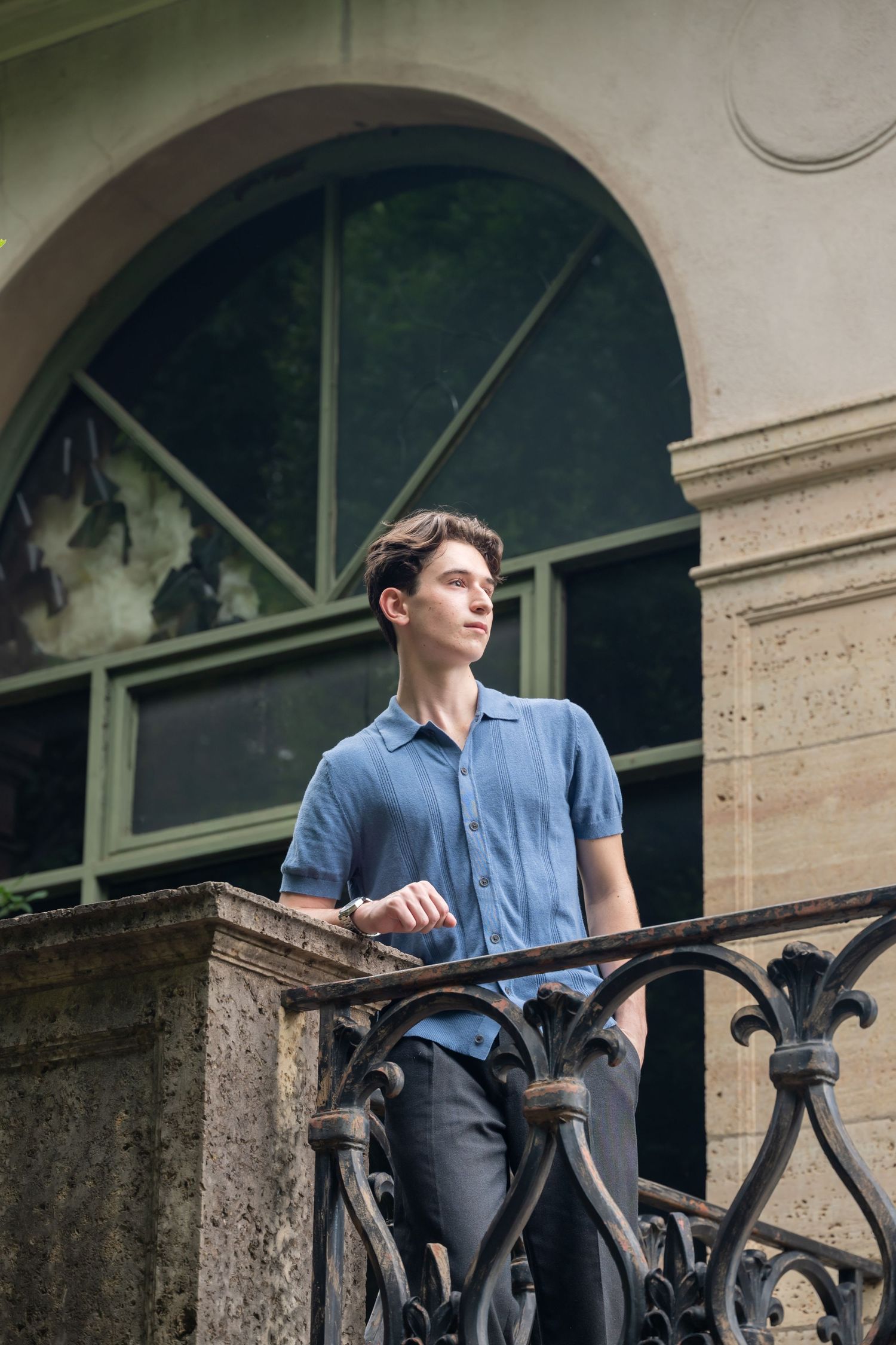 Young man in blue shirt leaning on ornate iron railing near arched window of historic building.