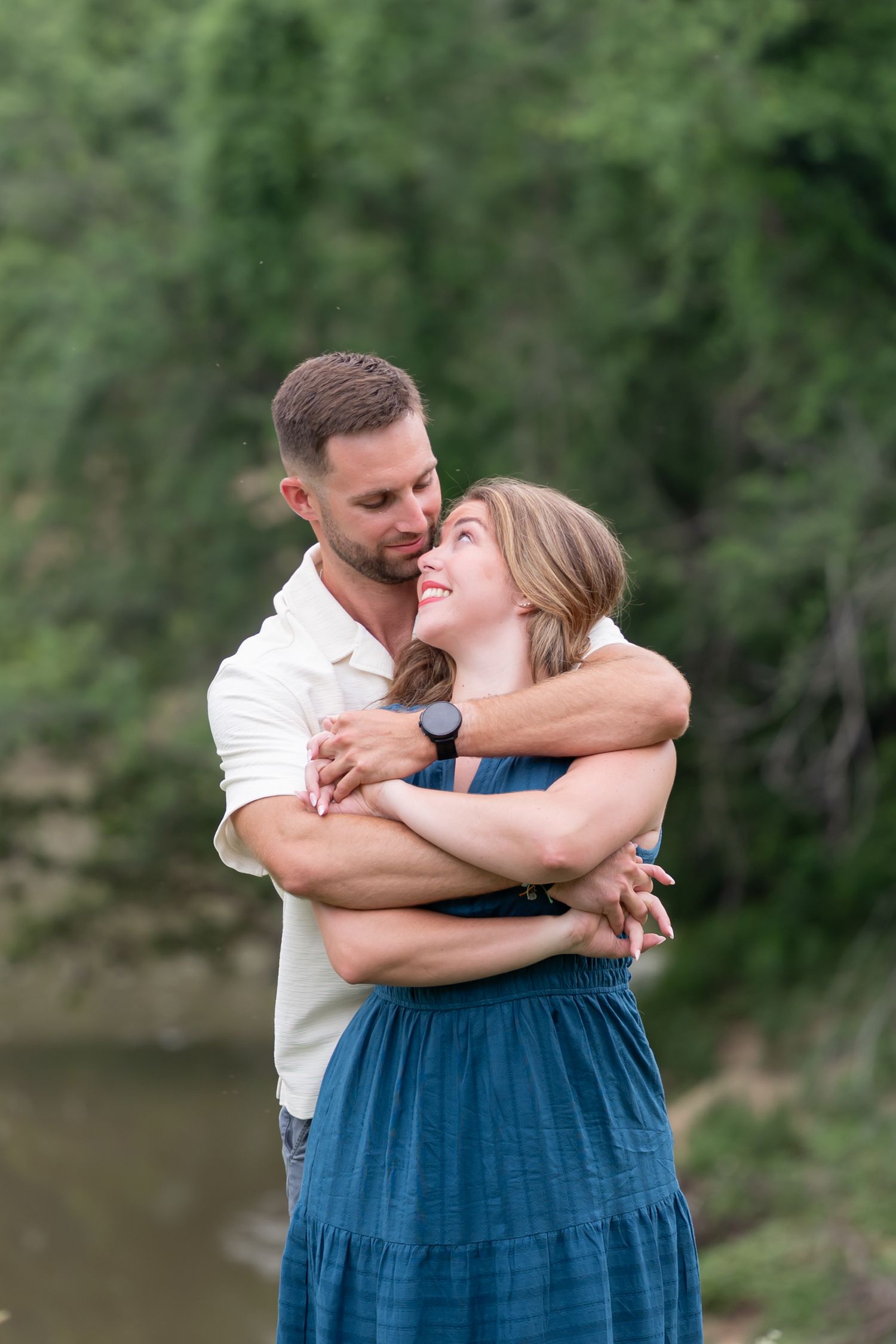 A couple embraces outdoors near a river, the man wrapping his arms around the woman in a blue dress.