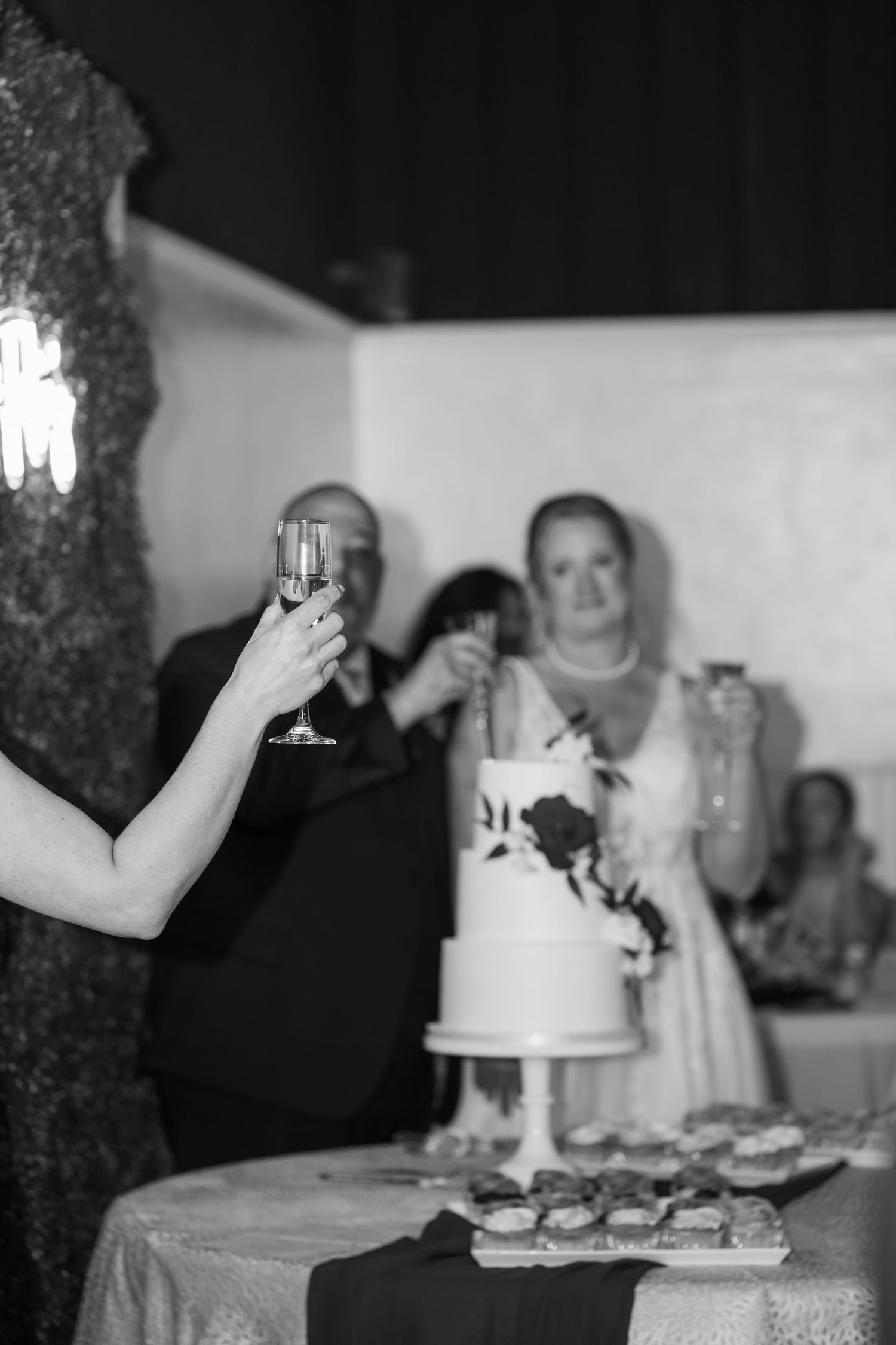 Black and white wedding reception photo with guests toasting champagne glasses near a decorated two-tier cake.