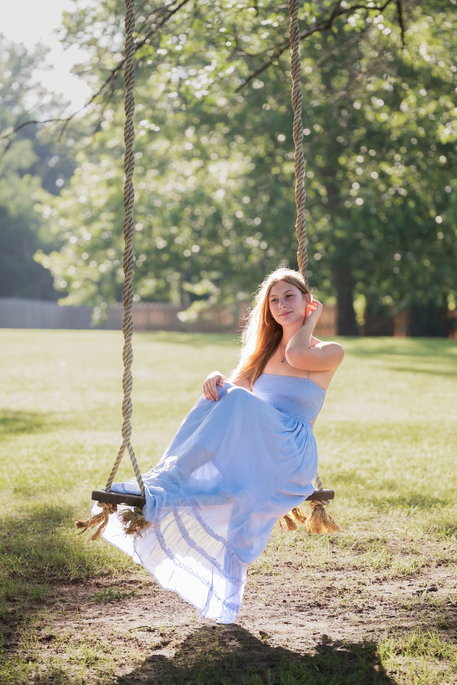 Young woman in a light blue strapless gown sitting on a rope swing in a sunny green park.