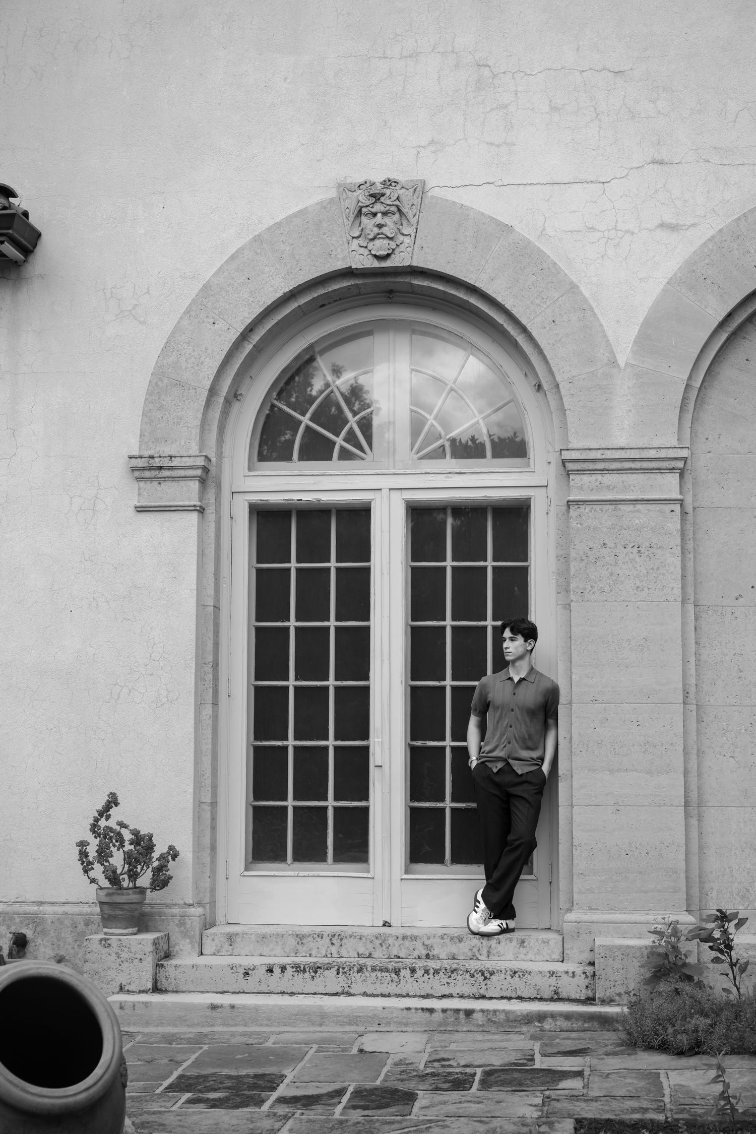 Young man leaning against ornate arched French doors of a classic stone building in black and white photo.