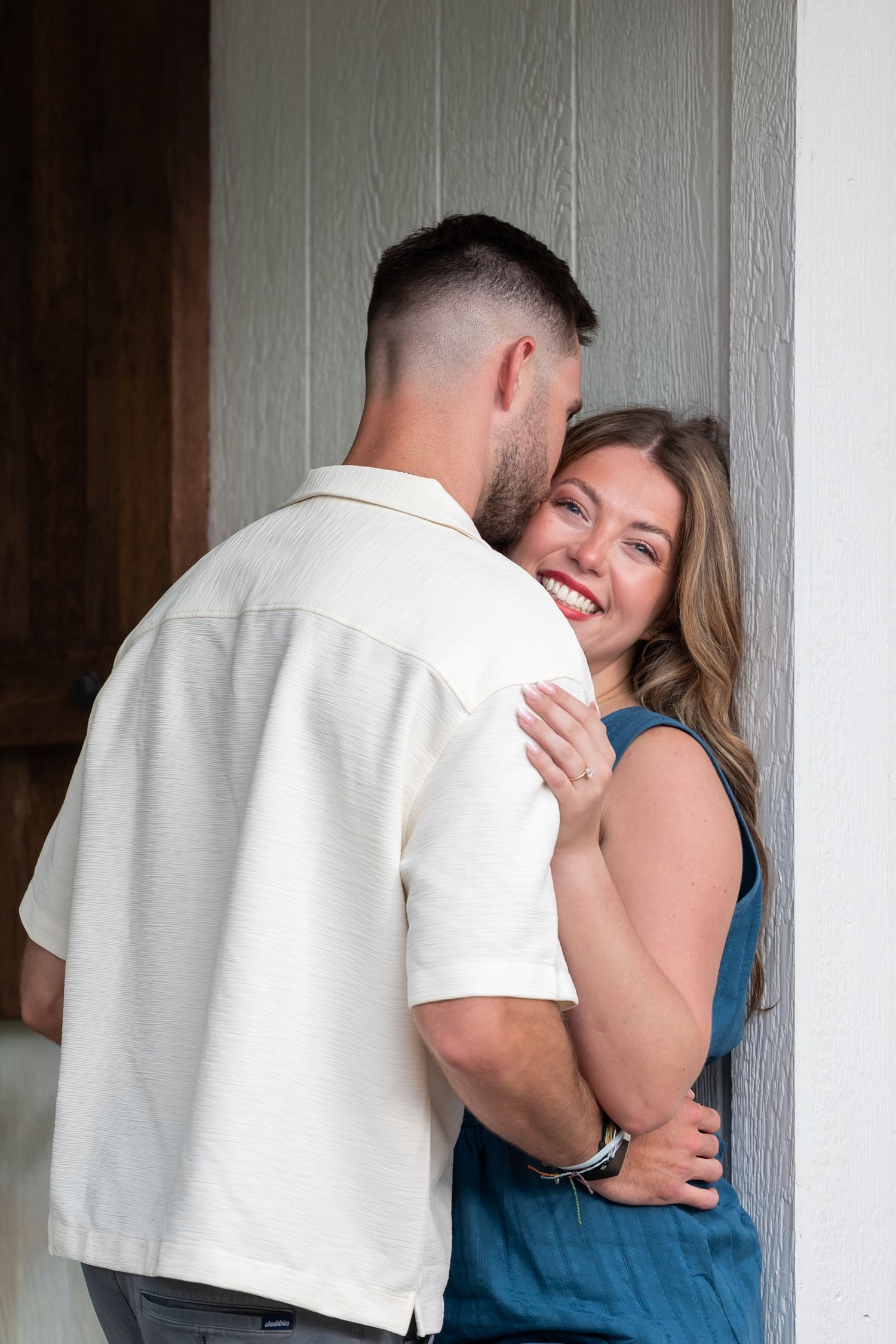 A smiling woman in a blue dress embraces a man in a white shirt against a gray wall outdoors.