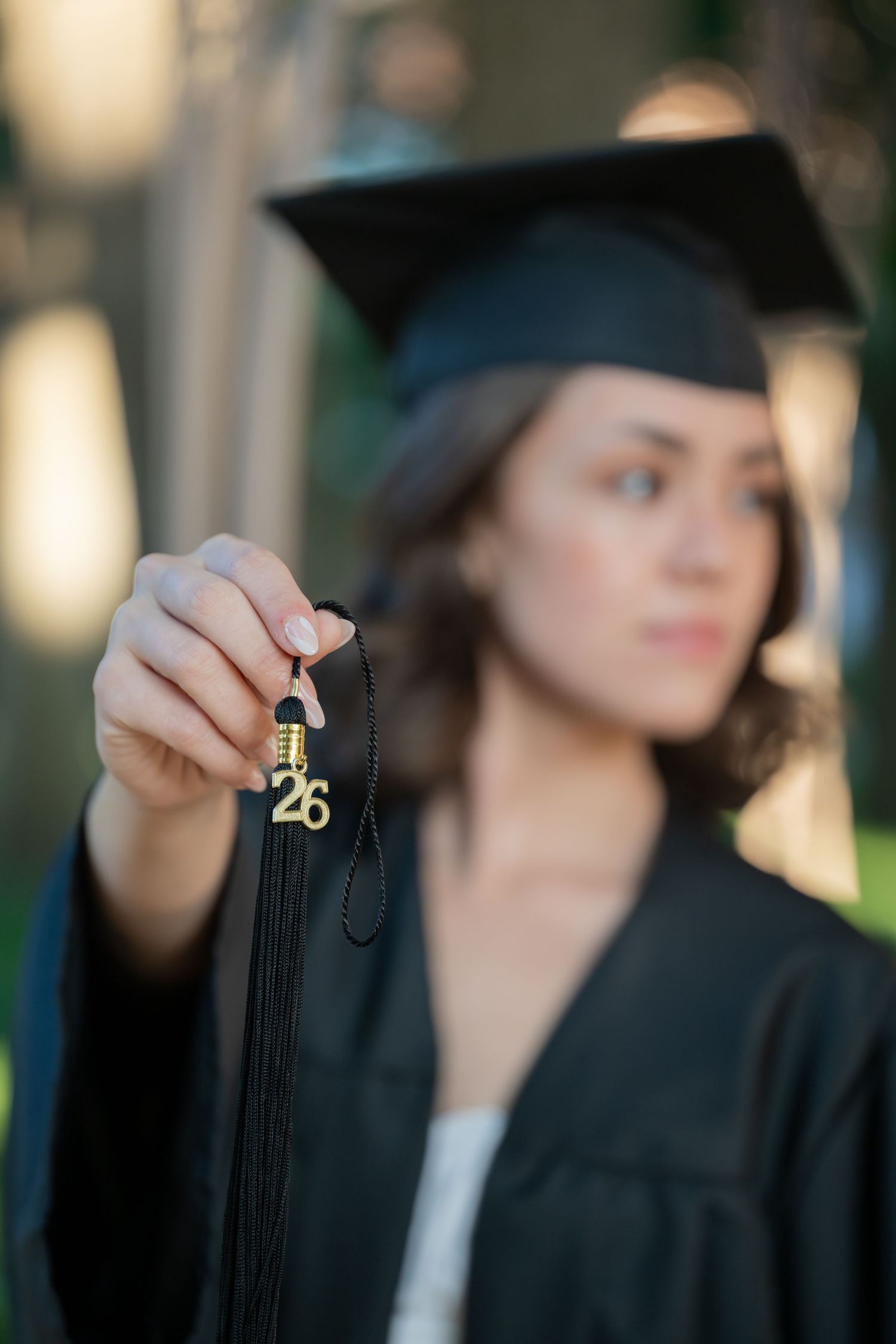 Graduate in black cap and gown holds up a gold 2026 tassel charm toward the camera outdoors.