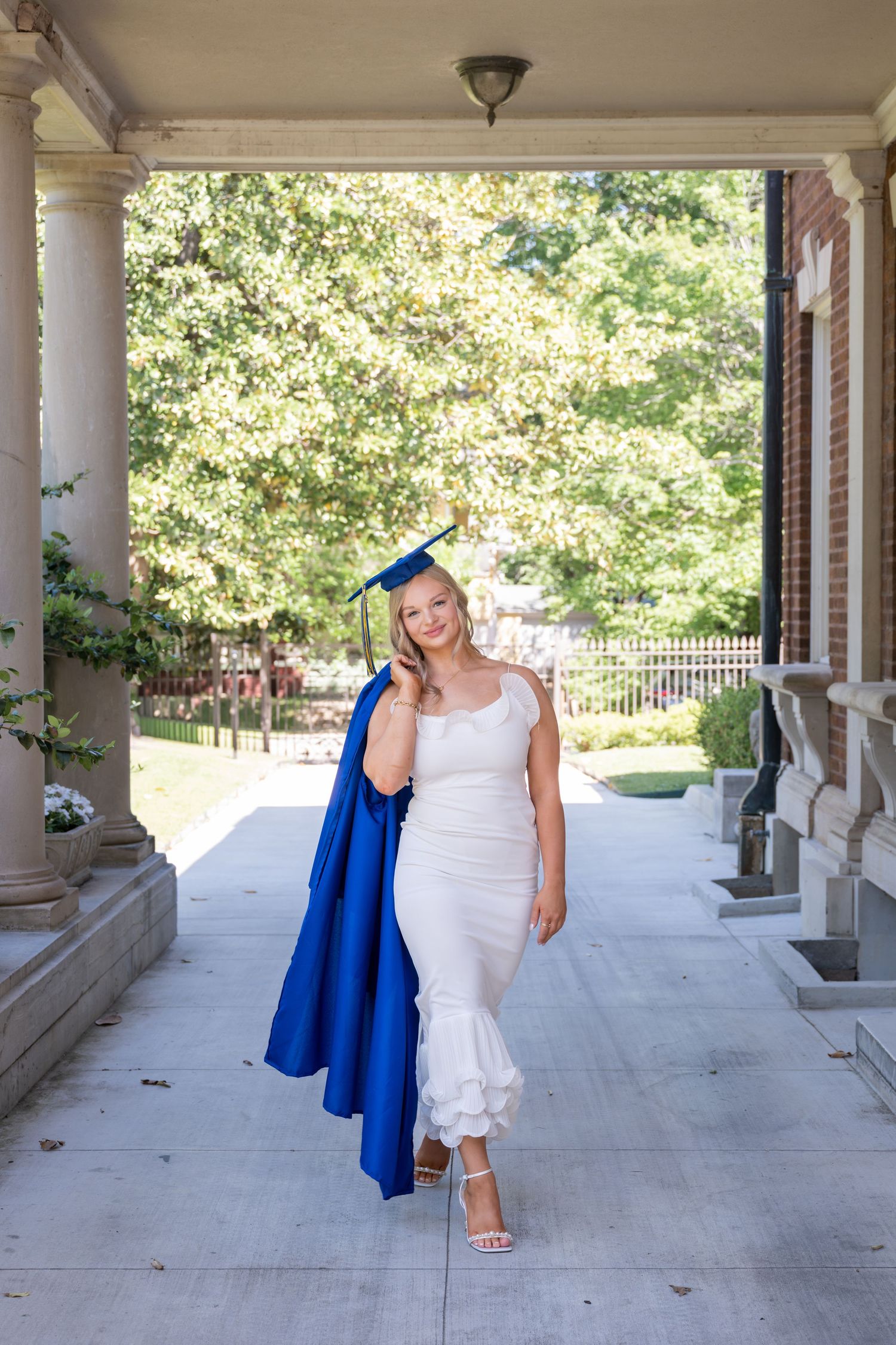 Young woman in white dress and blue graduation cap and gown posing on a college campus walkway.