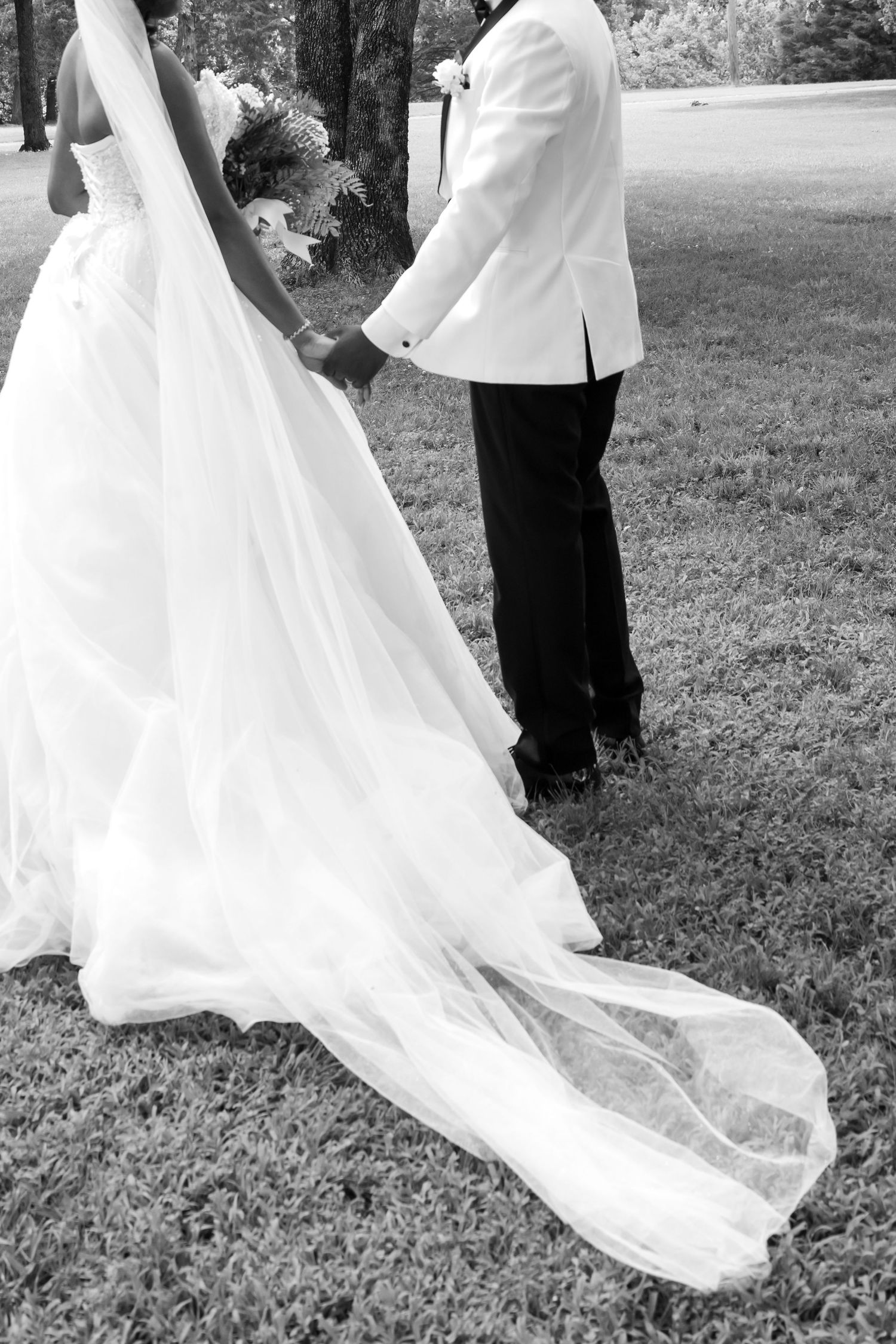 Black and white photo of bride and groom holding hands outdoors, bride wearing flowing white gown with long train.
