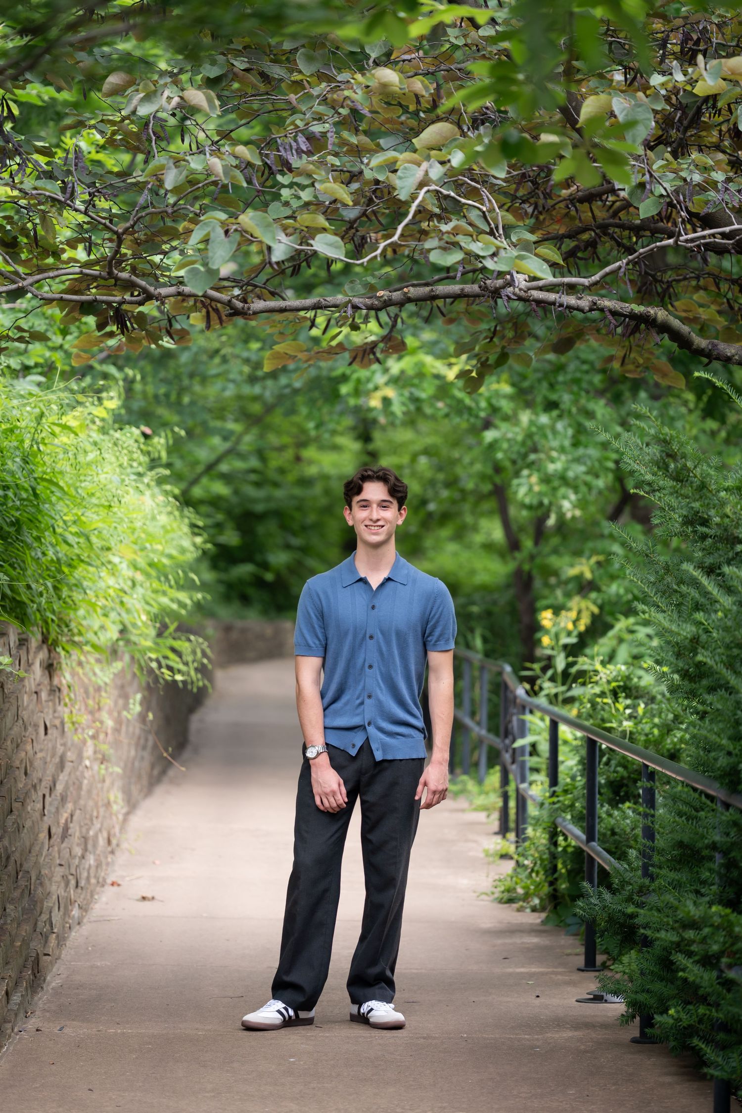 Smiling young man standing on a tree-lined pathway surrounded by lush green foliage.